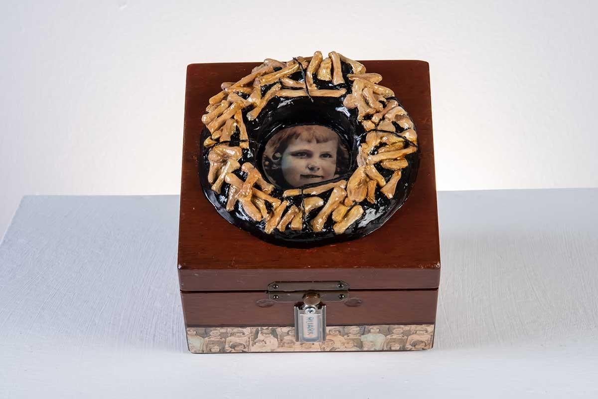 'Ruark', by Sunni Mercer is a sculptural mixed media wooden box measuring at 2 x 5.5 x 3.75 inches. Sunni uses found objects and photos in addition to the dark brown wooden box in her sculpture. There is a circular photo of a little girl which