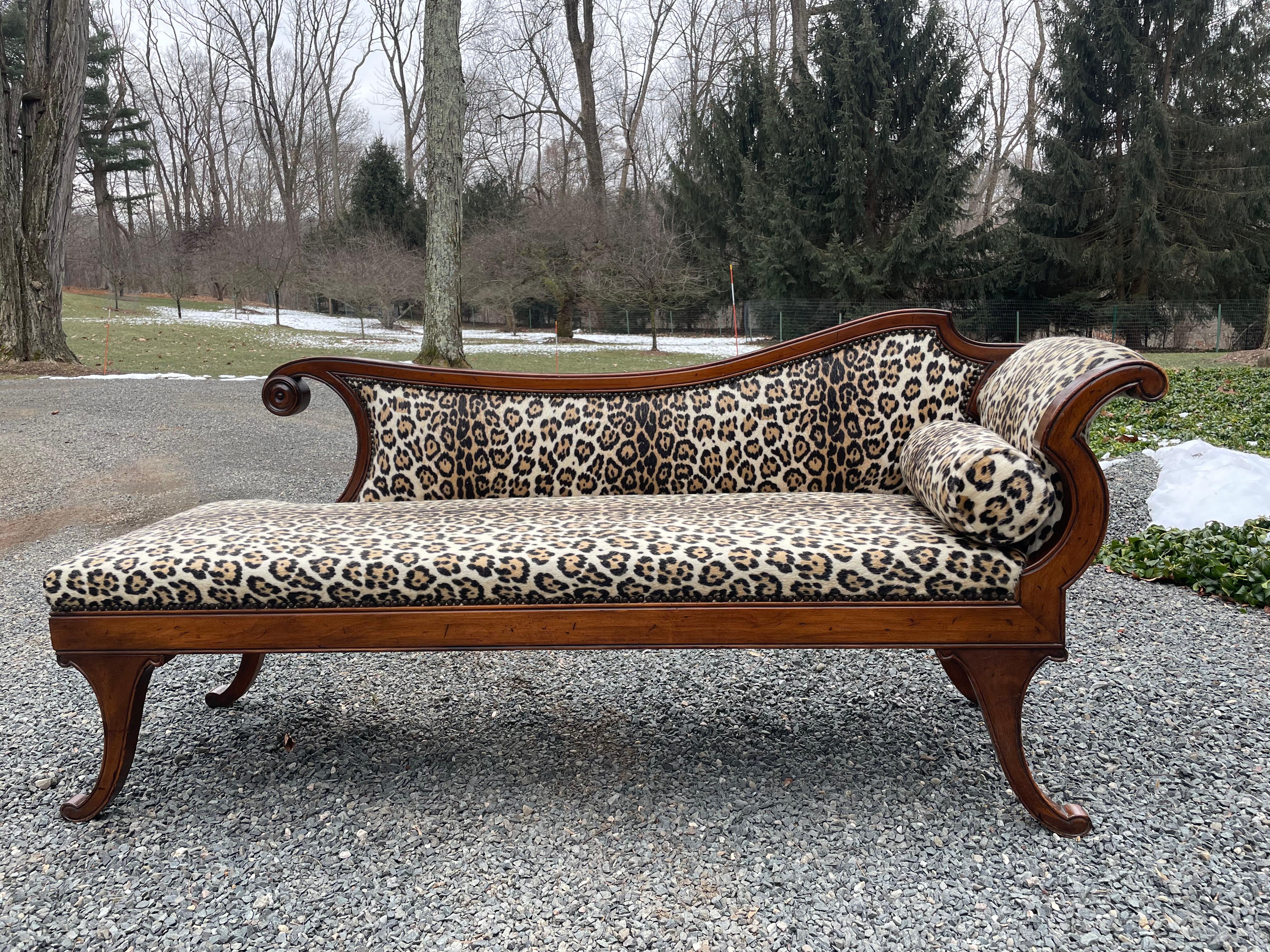 A super glam vintage Regency style chaise lounge or recamier upholstered in a timeless dramatic leopard faux fur. The wood has a cherry finish and the has nailhead detailing. Rolled cushion included.