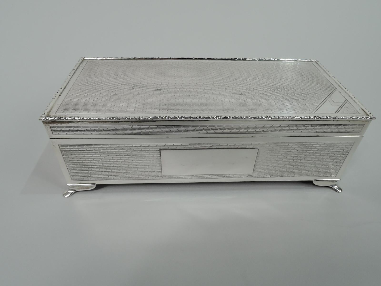 Art Deco sterling silver box. Made by Harman Bros in Birmingham in 1990. Rectangular with straight sides. Cover flat and hinged. Corner bracket supports. Engine-turned ornament in plain frames on sides and cover top. Front side has applied