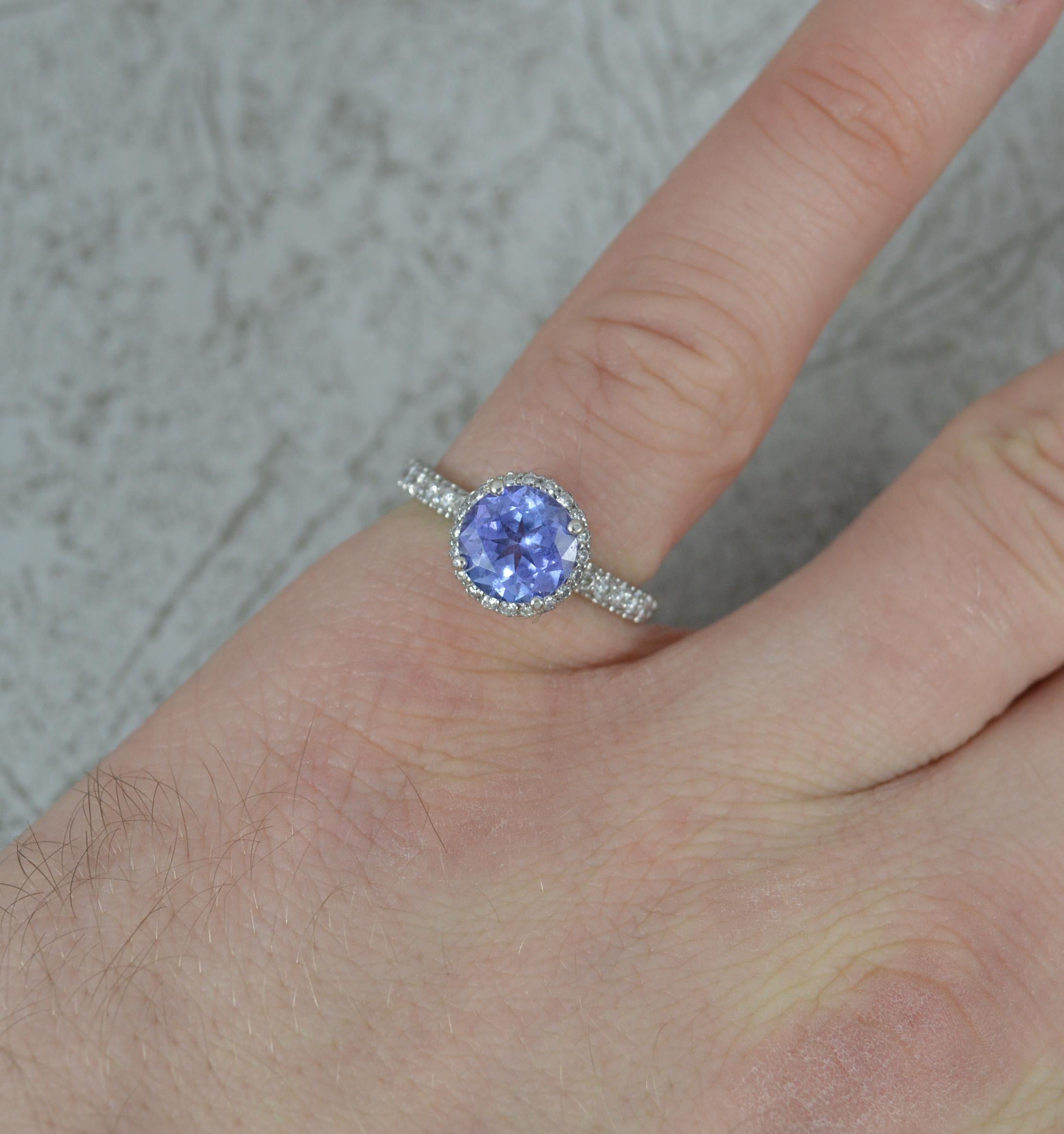 A wonderful halo engagement ring.
Solid 18 carat white gold example. Full of engraving throughout.
Set with a round cut tanzanite to centre, 7.4mm diameter. 
Further round brilliant cut diamonds to the head and shoulders. To total 0.75 carats