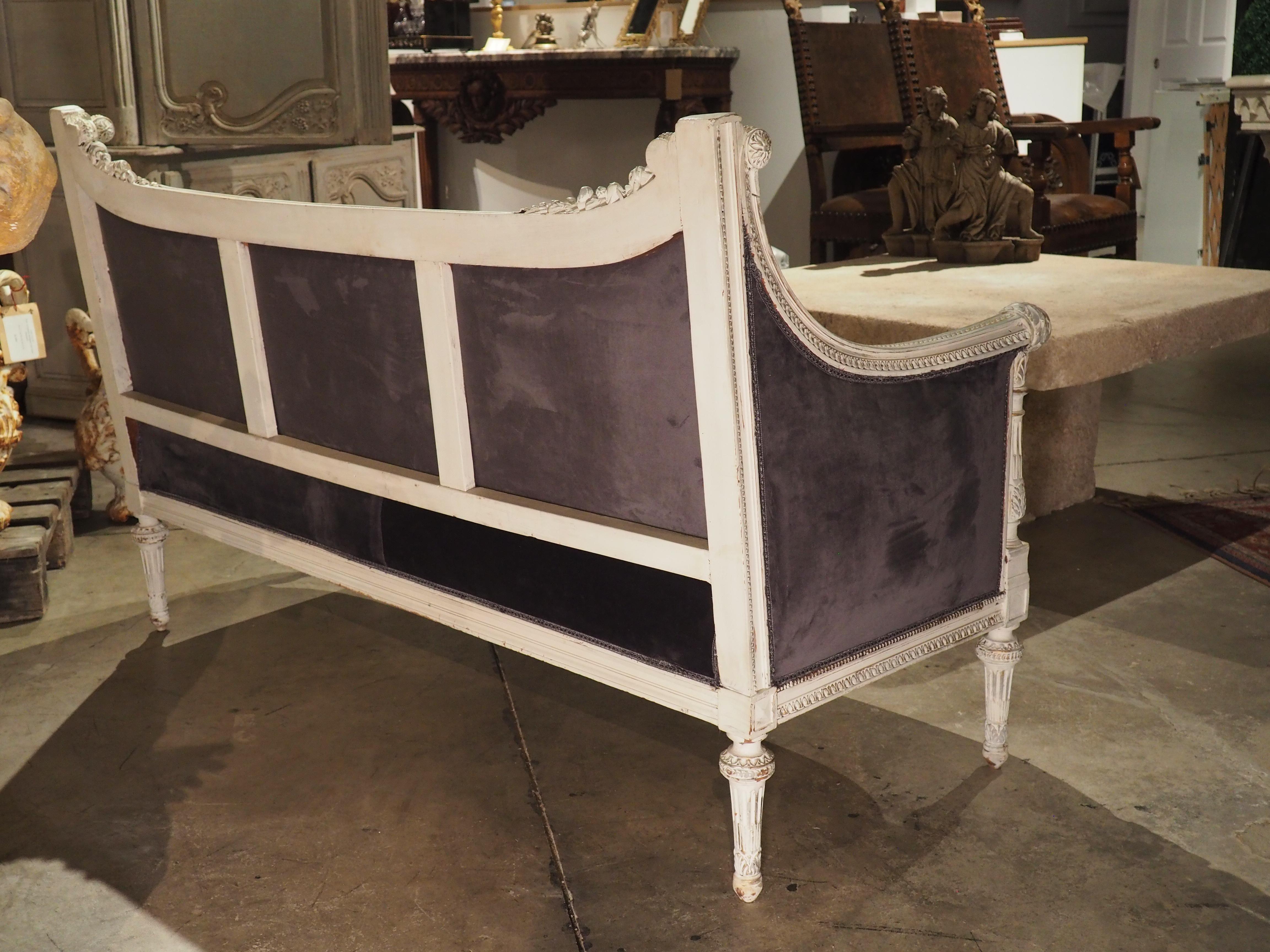 Hand-carved in the style of Louis XVI, this French canape features painted wood and a heather purple velvet upholstery with a gimp border. The highly detailed wood has been patinated in an antique white color with gold and silver rechampi subtly