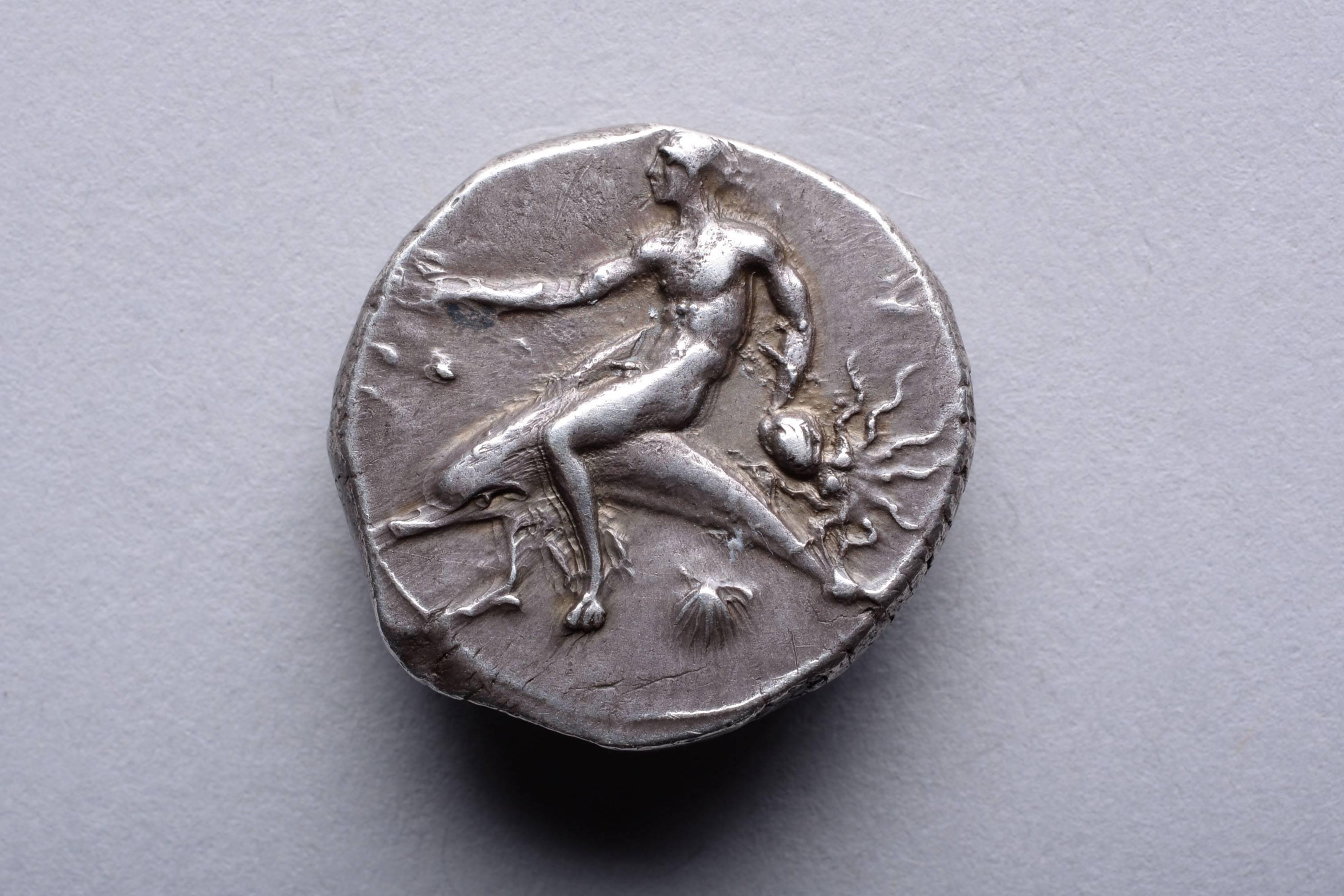 The obverse with the Greek hero Taras, nude and riding a dolphin, raising his right hand and clutching a struggling octopus in his left. The reverse with the hero seated on a low stool, swathed in thin drapery, a perfume flask dangling from a string