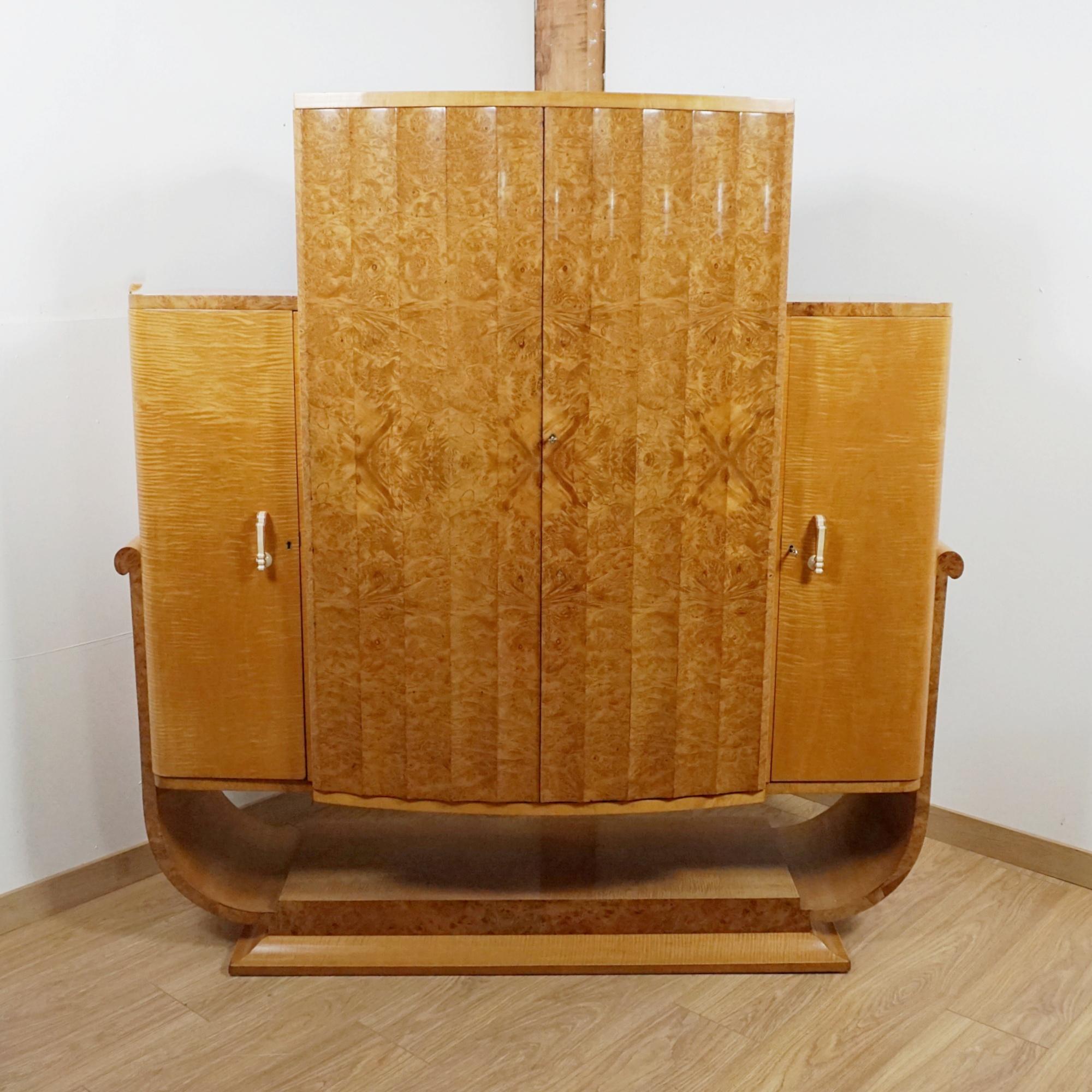 An Art Deco Cocktail Cabinet by Harry & Lou Epstein. Bur walnut veneered front fluted cabinet with satin birch veneer and banding. Central section opens to reveal a mirrored lit satin birch lined interior with lower pull out mixing tray, above a
