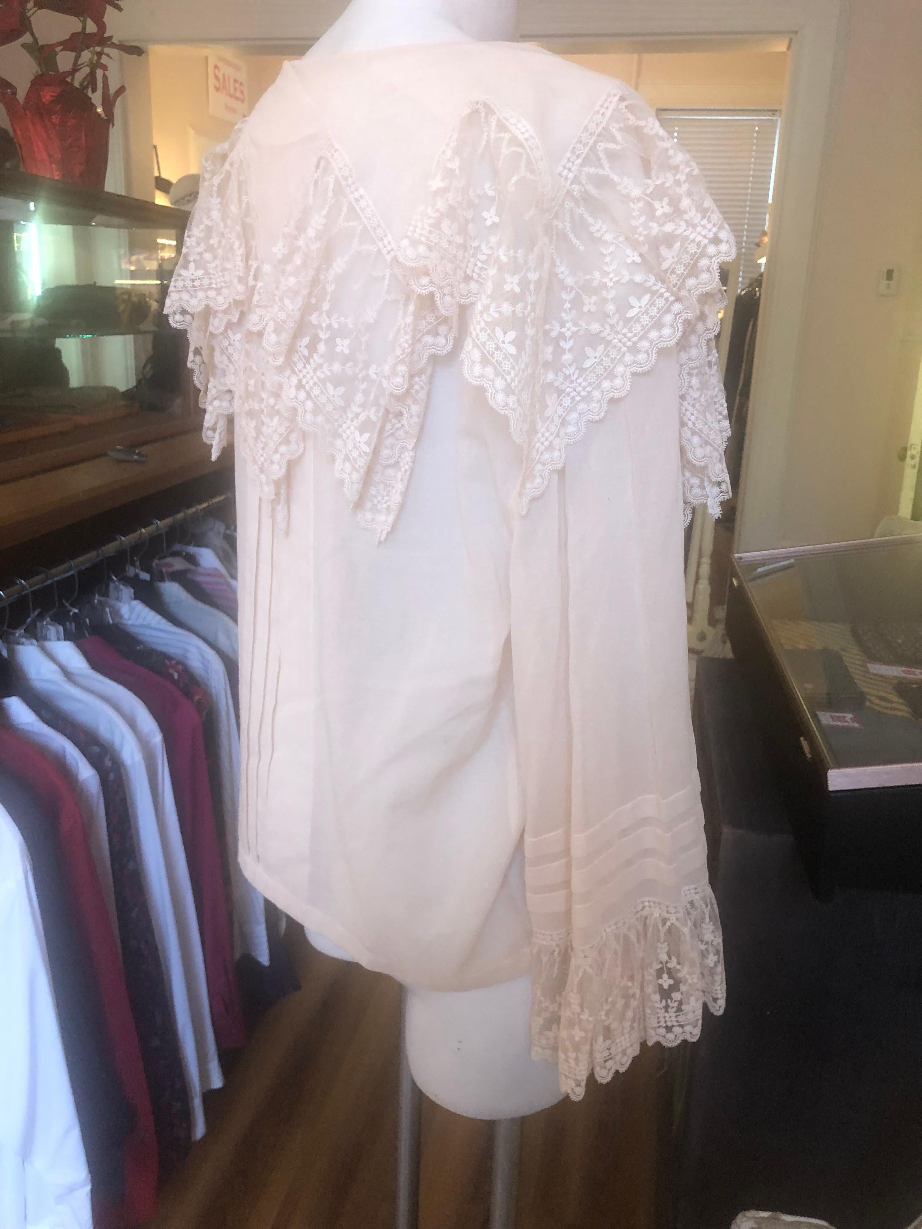 This is a spectacular never worn ecru cotton blouse with lace around the collar, front and cape like at the back. Lace is also present on the cuff of the wide sleeves. There are also permanent pleats on the back, front, and sleeves. The buttons are