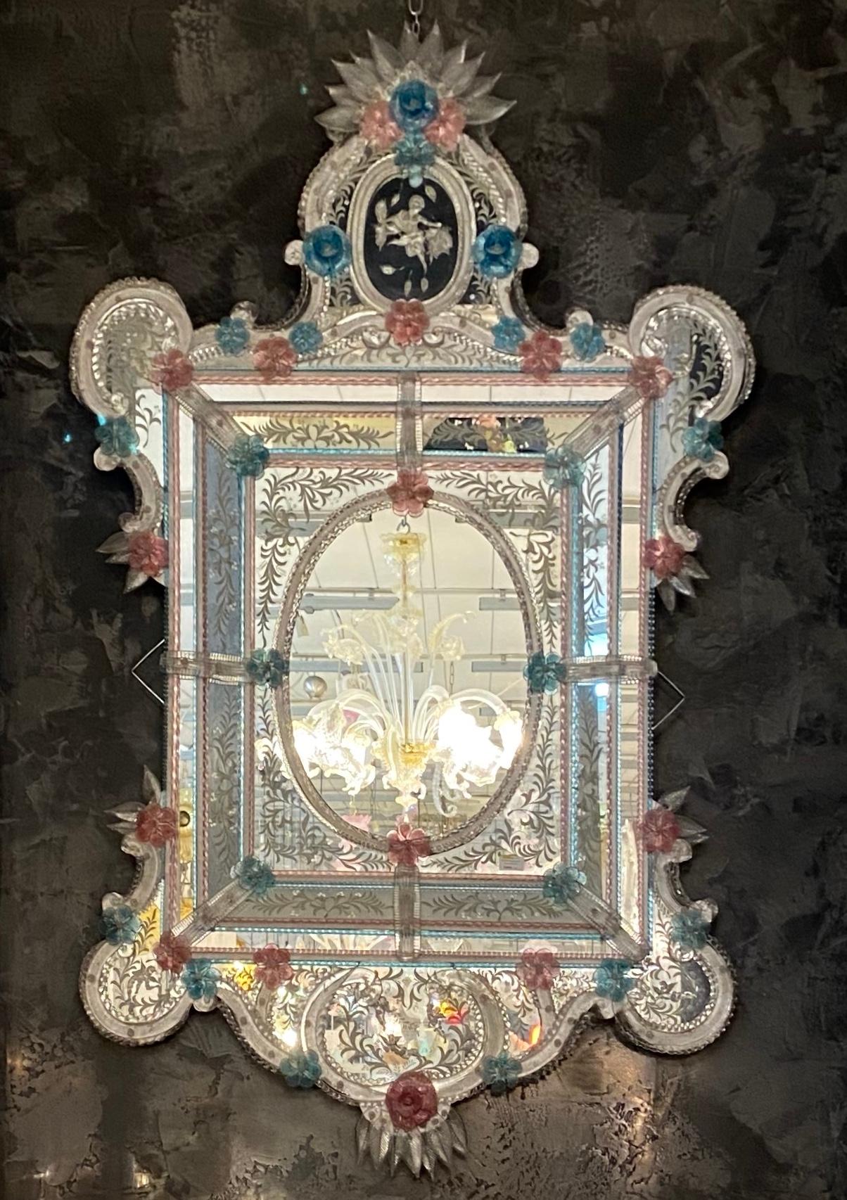 Dieser schöne venezianische Spiegel ist mit geätzten Blumenmotiven versehen, die den Spiegelrahmen zieren. Entlang der Ränder des Rahmens befinden sich gläserne Seilakzente und zahlreiche rosa und blaue Glasblumen.
Ausgeführt von einem großen