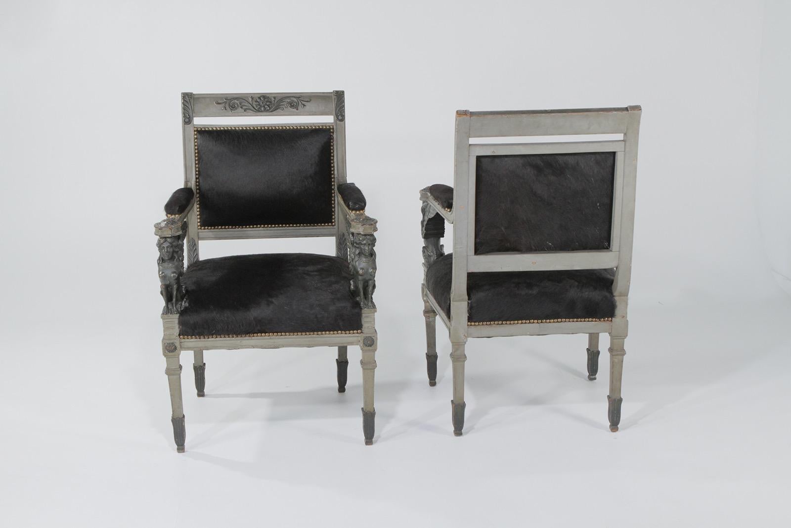Truly amazing ornately carved wood Egyptian Revival armchairs having a gilded underpaint with grey wash paint, glamorous details including rosettes and female sphinx figures on the arms, and glamorously contemporized with black cowhide upholstery.