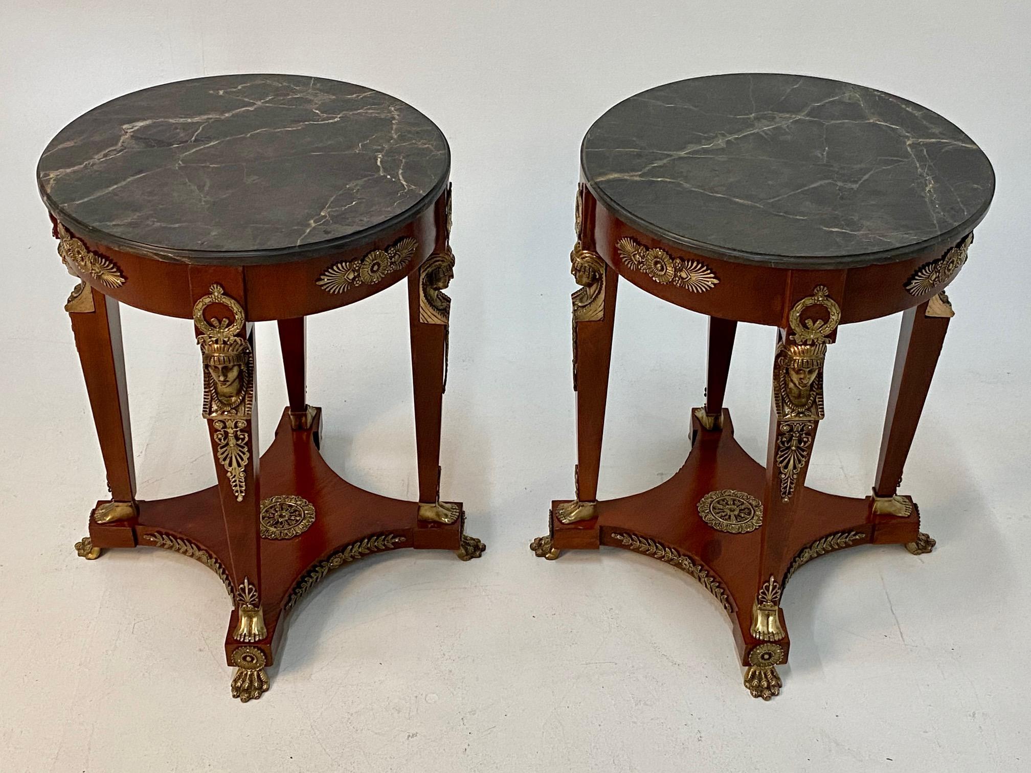 Elegant and ornate pair of round French Empire style end tables having rich mahogany bases with 4 legs and fancy bronze figural decoration at the tops of the legs, wreaths, and animal paw feet on elevated bases. The black and grey veined marble tops