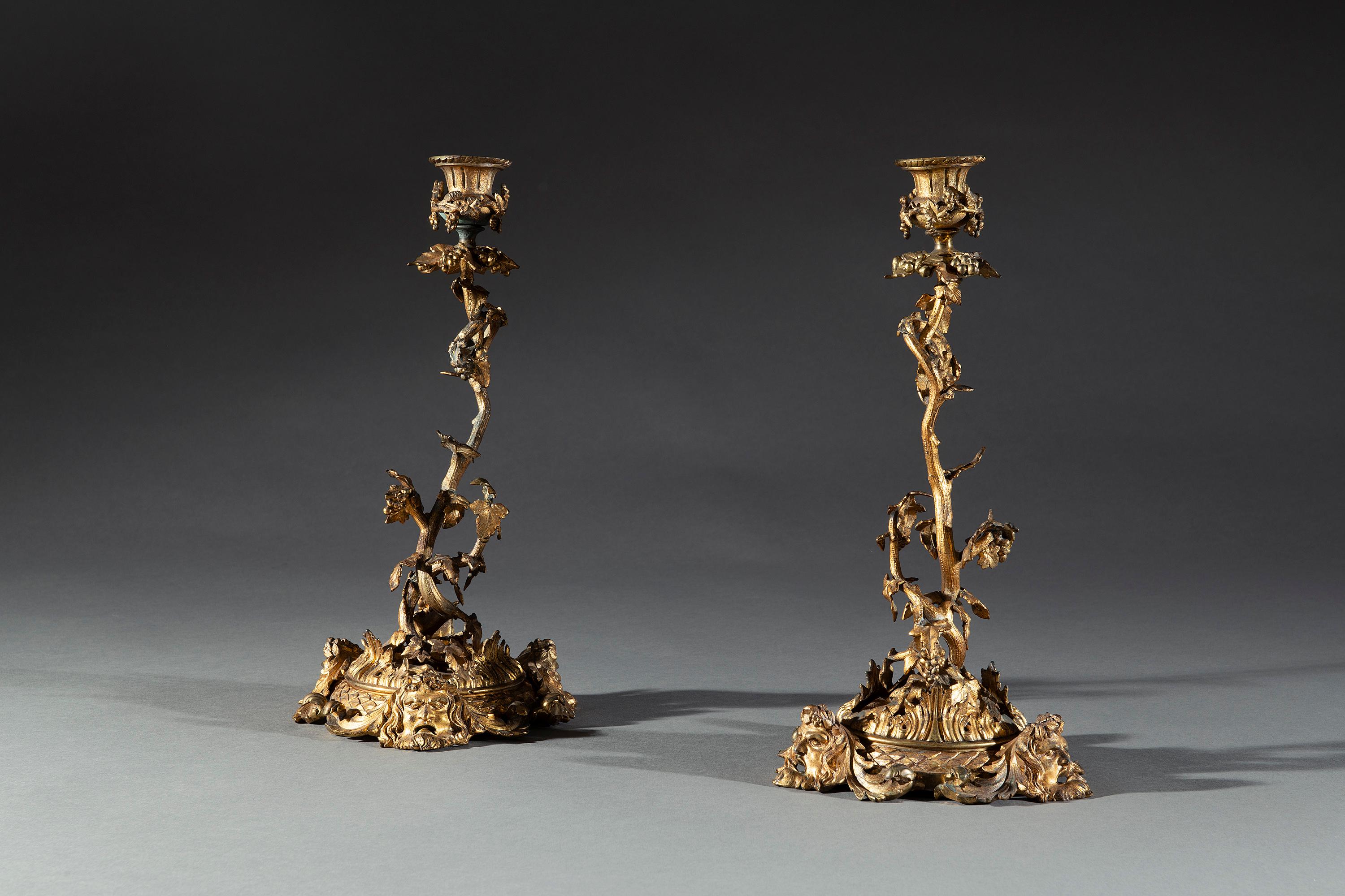 The large pair of gilt candlesticks with cast thistle cup candle holders are adorned with vine leaves and grapes. The stems each consist of a twisting vine with leaves and bunches of young grapes. The vine stems emerge from rising leaves and a