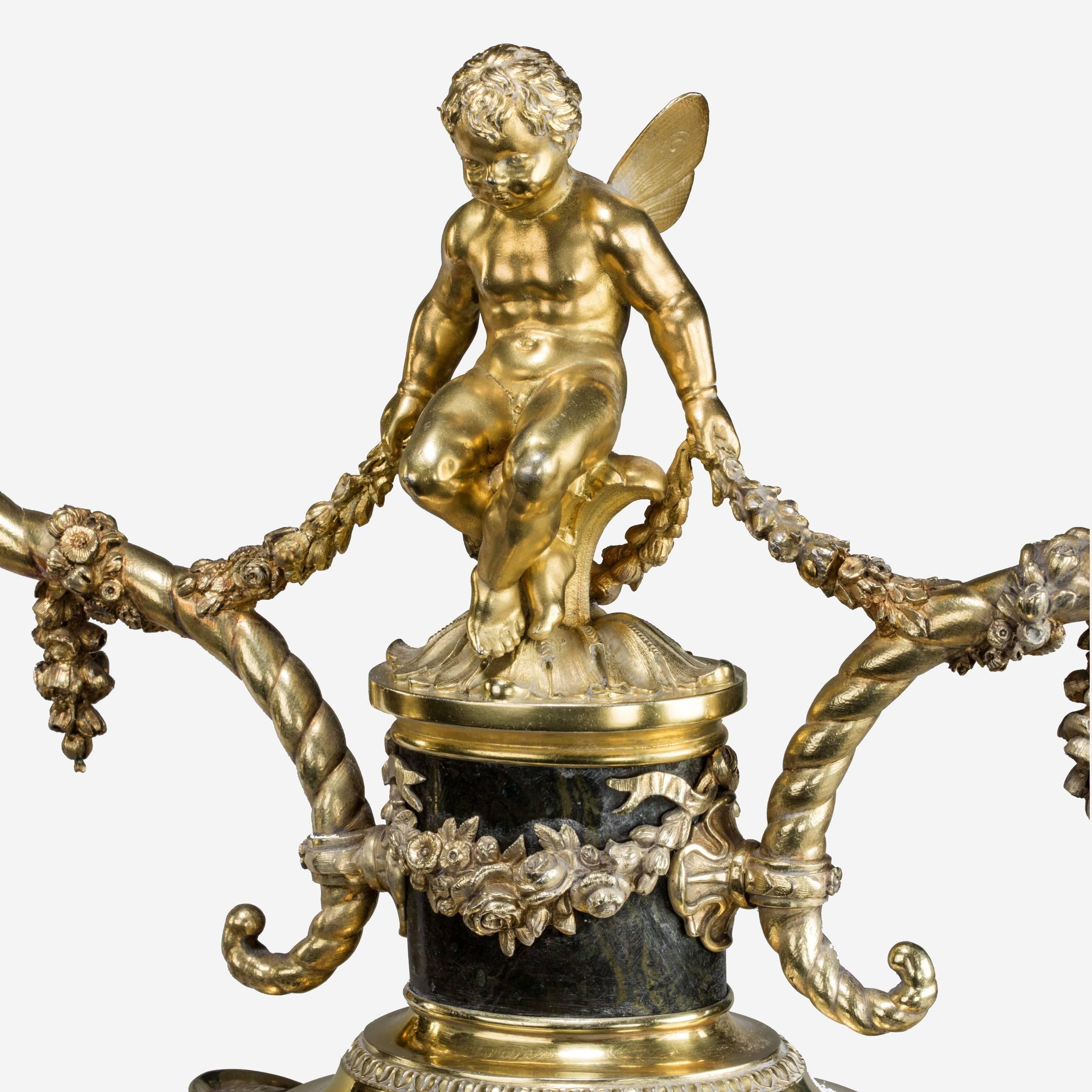 A superb pair of Victorian silver gilt candelabra,
each comprising a serpentine footed urn supporting a winged cherub sitting on two cornucopia festooned with floral garlands, applied with classical mounts in the form of Grecian goddess masks,