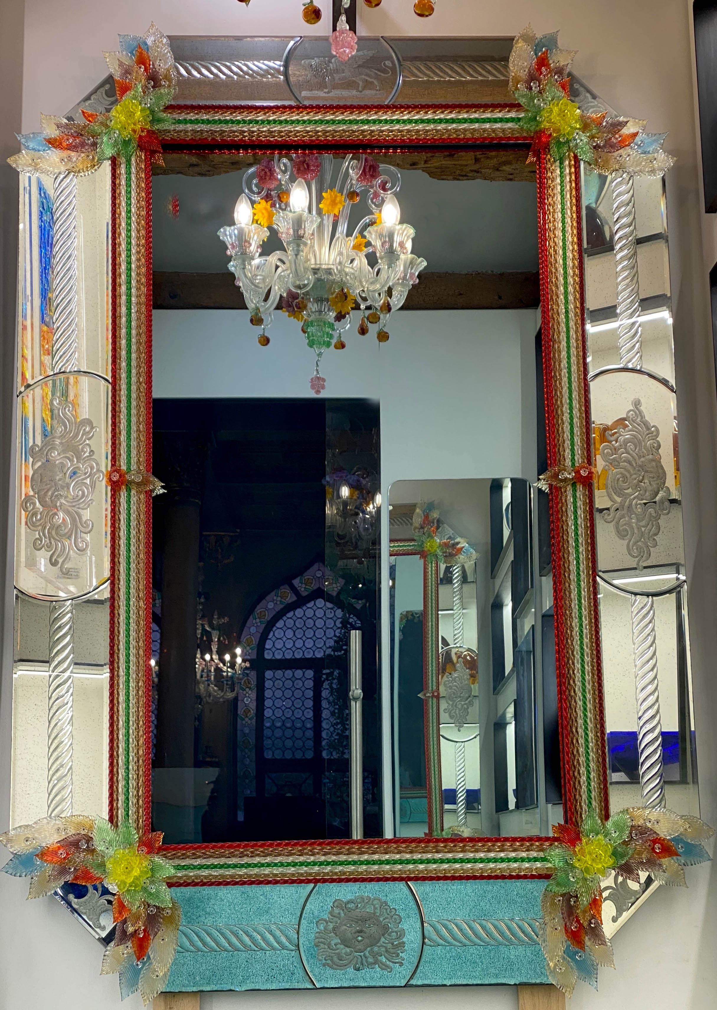 This beautiful Venetian mirror features etched figure motifs adorning the mirrored frame. Centered by a lion figure symbol of Venice. Along the edges of the frame are colorful glass rope accents and numerous multicolor glass leaves and