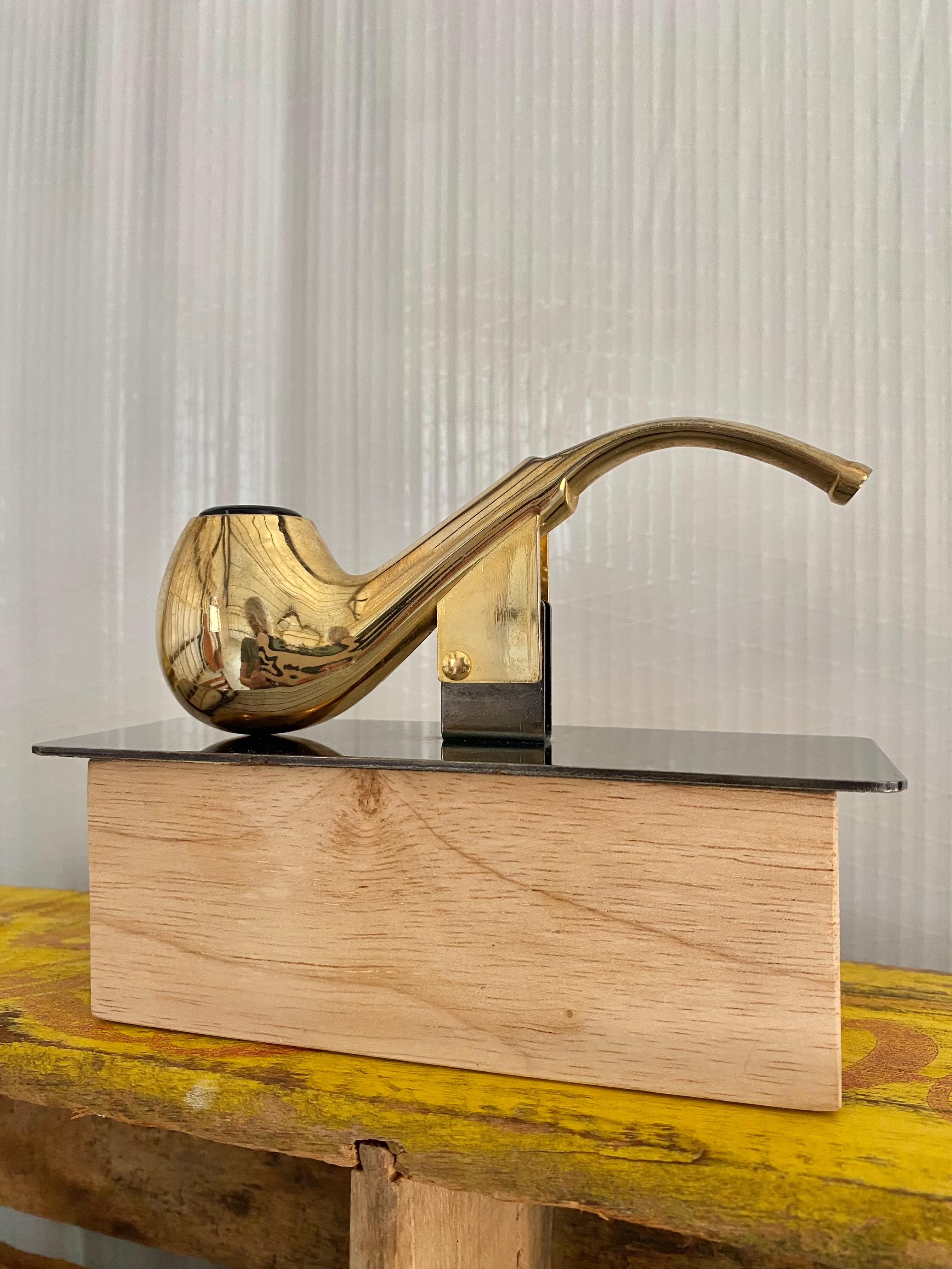 Una pipa surrealista in ottone di peso elevato, caricata a molla per funzionare come porta banconote e con un'apertura per contenere una penna. 

La base in metallo piatto bronzato è fissata al tubo e una base in legno separata è inclusa per