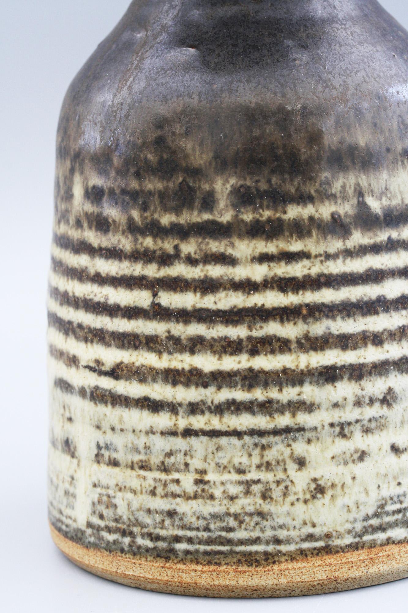 A stylish thrown and glazed stoneware studio pottery vase with a ribbed body attributed to Susan Bennett (b.1954) and dated 1977. The vase has a bell shaped body with a wide flat rounded and unglazed base and has a pinched neck and large trumpet