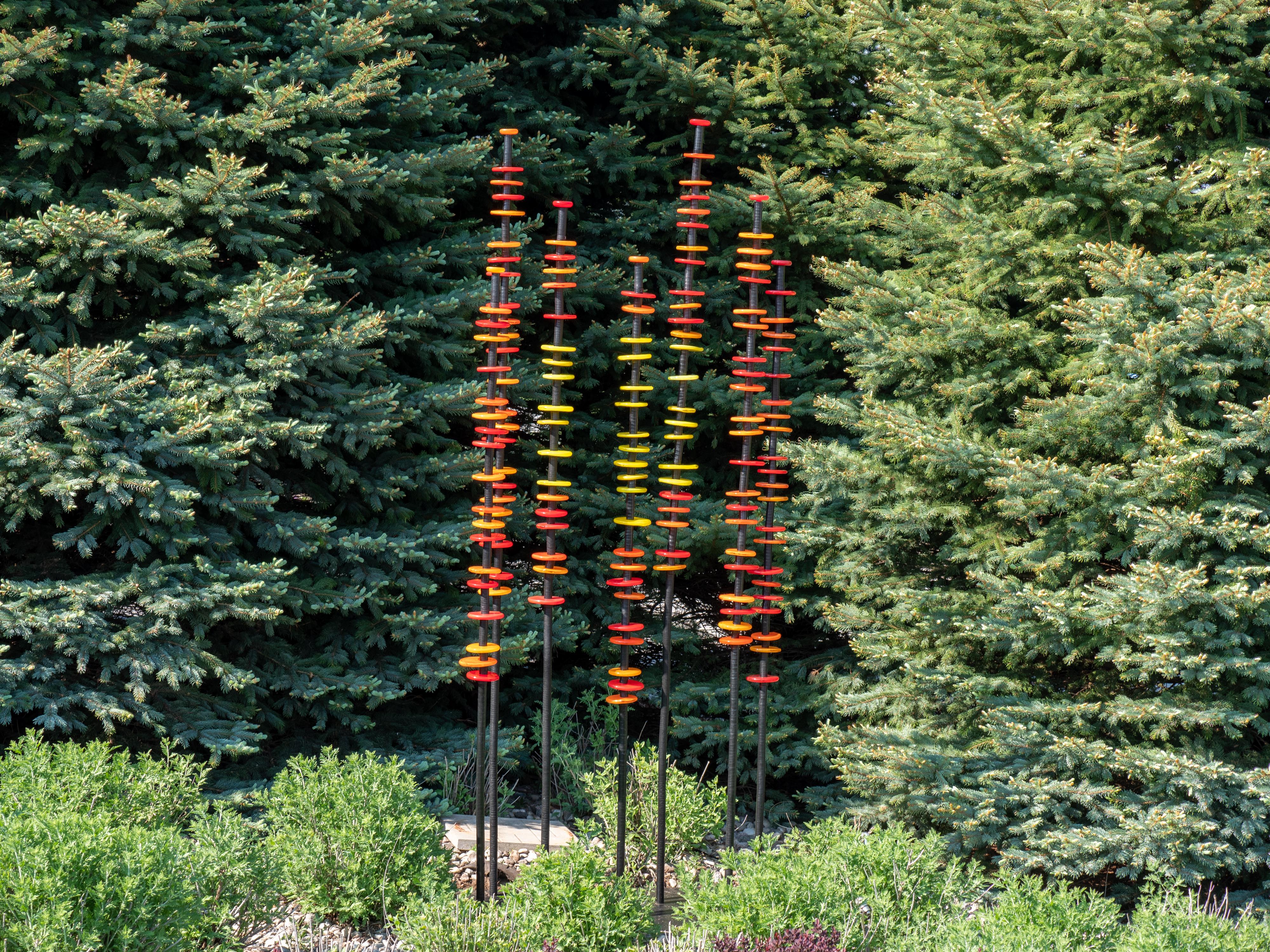 Hand blown glass rings of bright red, orange and yellow are stacked organically on seven stems in this brilliant outdoor sculpture by Susan Rankin. The stems are fixed to a steel plate and sway gently in the breeze.  Each column includes the hand
