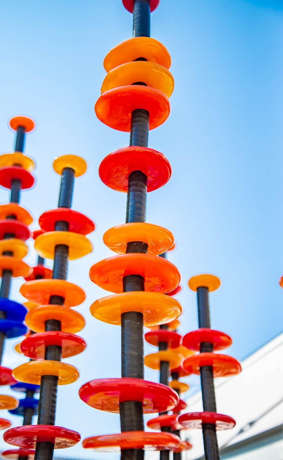 Hand blown glass rings of bright red, orange, yellow and blue are stacked organically on ten stems in this brilliant outdoor sculpture by Susan Rankin. The stems are fixed to a steel plate and sway gently in the breeze. Each column includes the hand