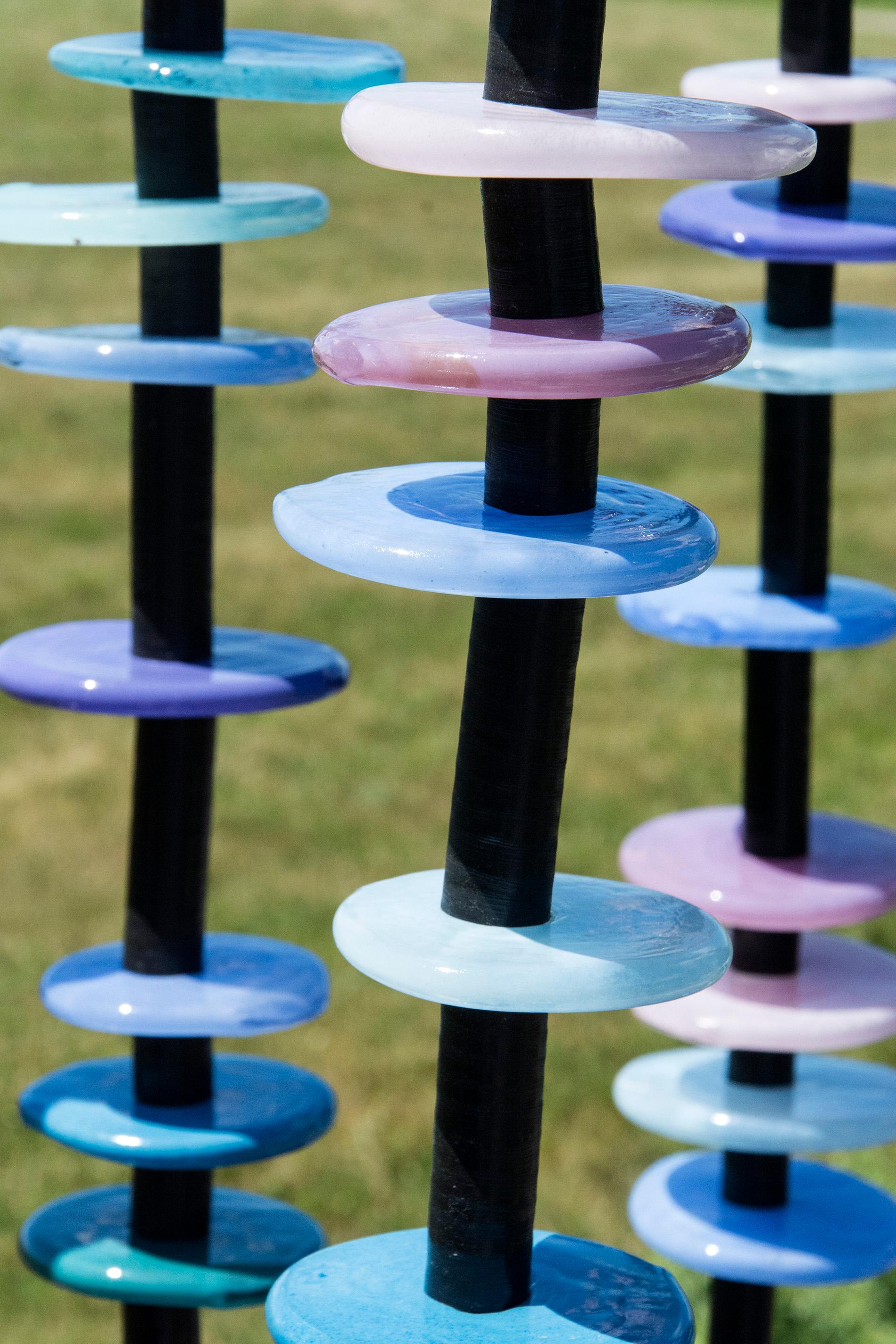 Hand blown glass rings of purple, violet and blue are stacked organically on five stems in this delightful outdoor sculpture by Susan Rankin. The stems are fixed to a steel plate and sway gently in the breeze.  Each column includes the hand formed