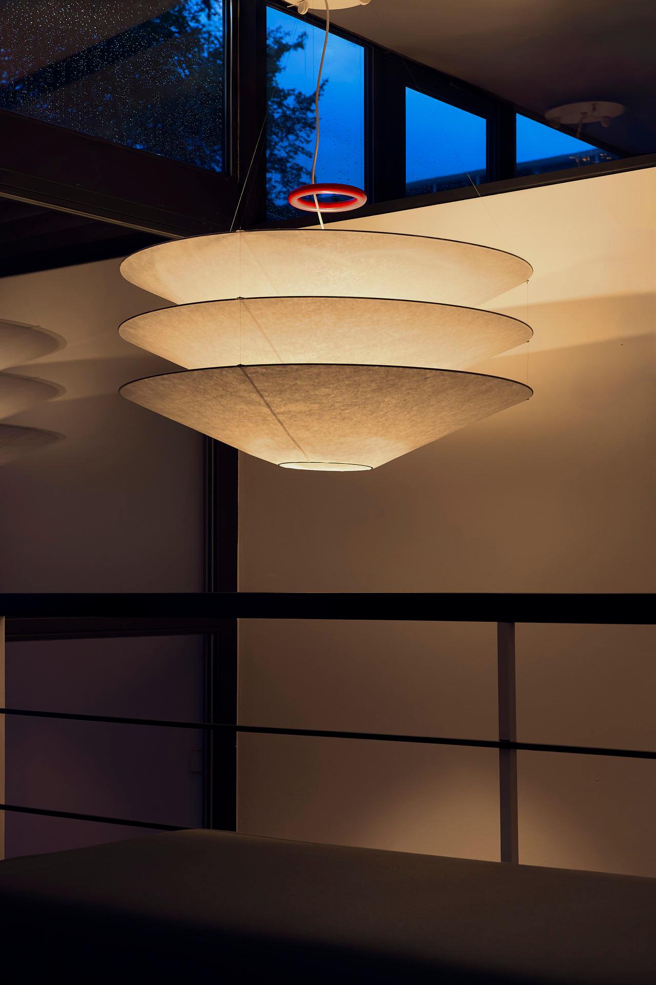 Floatation – Version 1
Designer: Ingo Maurer (1980)

Floatation is a delicate pendant lamp made of Japanese paper and metal. The red iron ring allows height adjustment, and the ceiling rose is finished in white. Handcrafted Japanese paper shades