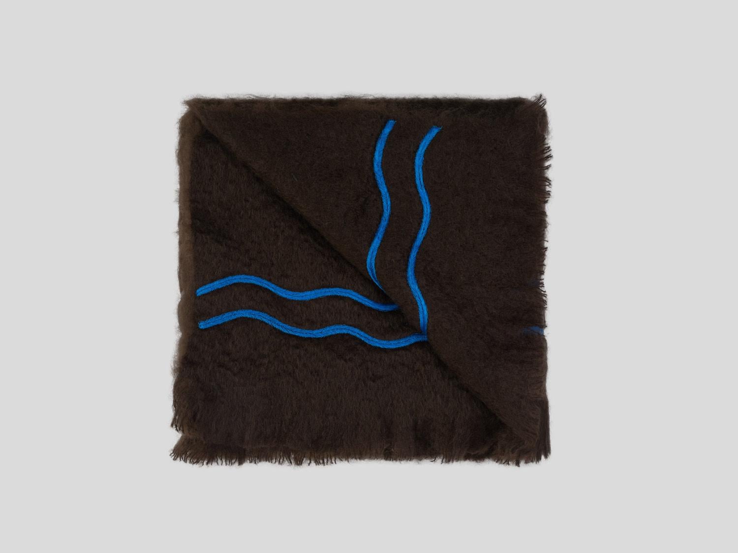 Suter, eine grafische braune Decke aus feinstem neuseeländischem Mohair. Diese Decke ist mit leuchtend blauen Stickereien verziert, die von Hand ausgeführt werden. Nichts ist so persönlich wie Ihr eigenes Interieur, die Farben des Stoffes und der