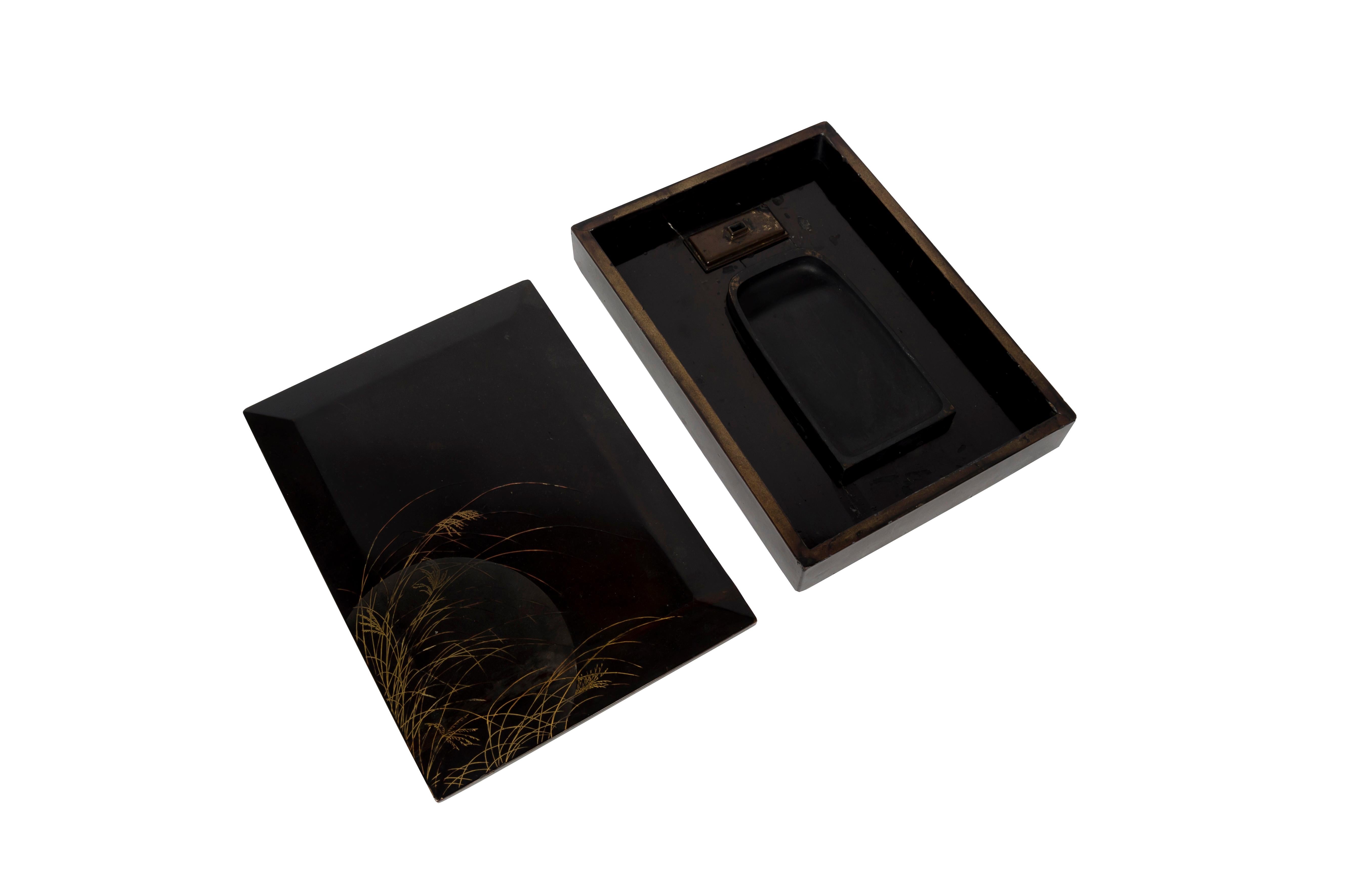 Ein altes Suzuribako oder Schreibkästchen aus schwarzem Lack mit facettiertem Deckel, das als einzige Verzierung im unteren linken Bereich eine aus hohen Gräsern auftauchende Mondscheinszene zeigt. Der Mond wird durch die Zugabe von Metallsalzen wie