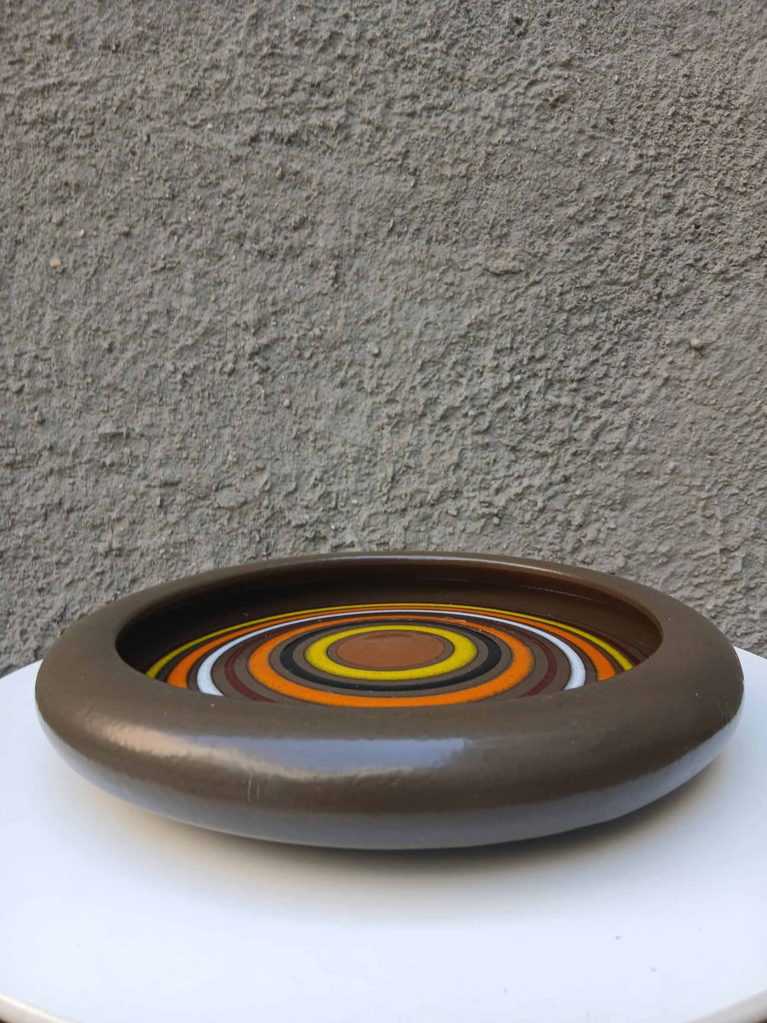 Fascinante pieza/posacabezas de cerámica de los años 70 con decoración Op-Art de cercos concentrados en tonos bronce, azafrán, giallo, blanco y negro. La forma acanalada con amplio bordo estrellado evidencia el diseño gráfico central, típico de la