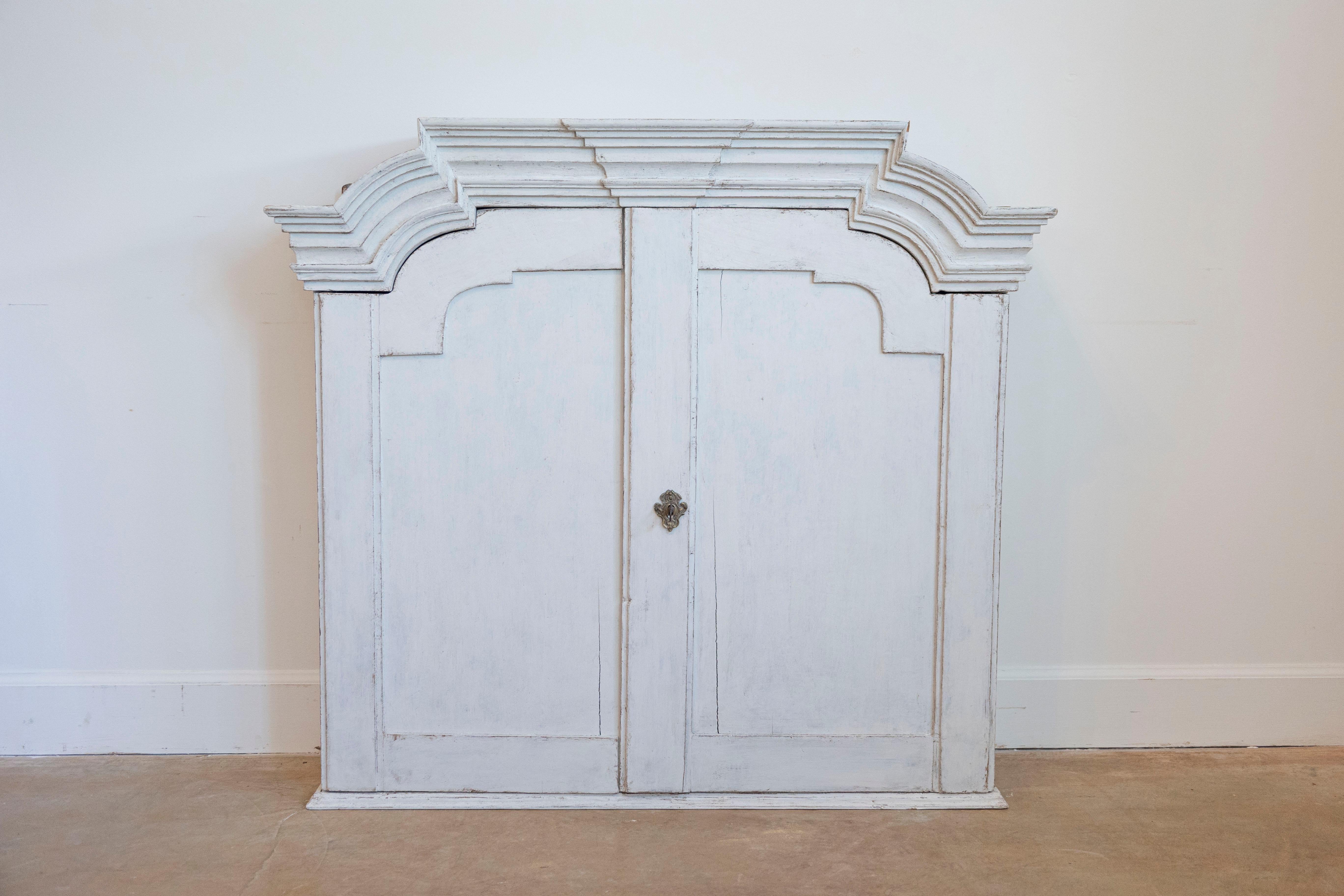 A Swedish Rococo period painted wood wall cabinet from the late 18th century, with chapeau de gendarme style cornice, two doors and three inner drawers. Created in Sweden during the third quarter of the 18th century, this Rococo period wall cabinet