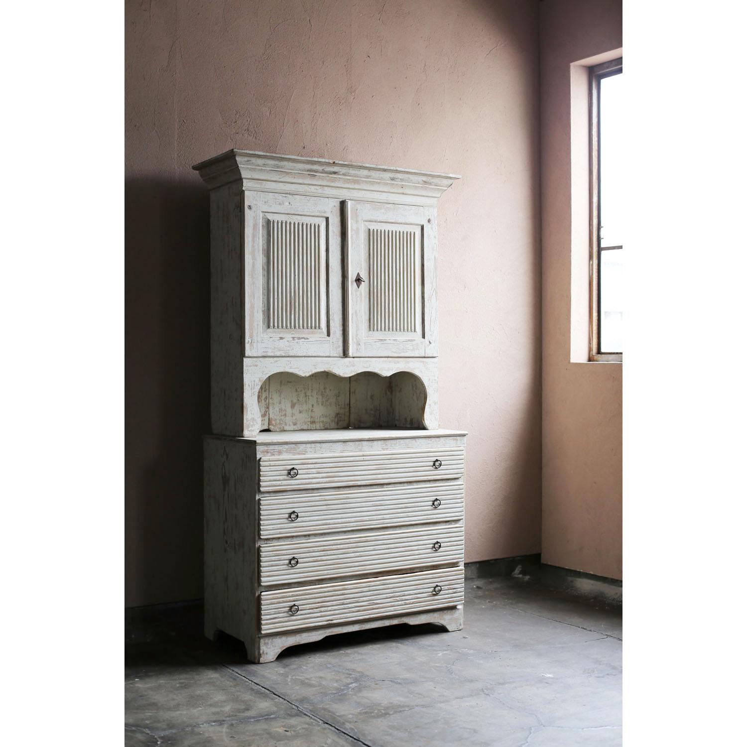 A two-tiered wooden cabinet from the early 19th century Swedish Gustavian period. The top section opens to a shelf for hanging wine glasses and a small drawer for storing cutlery. The middle section has an open shelf with scalloped accents. The