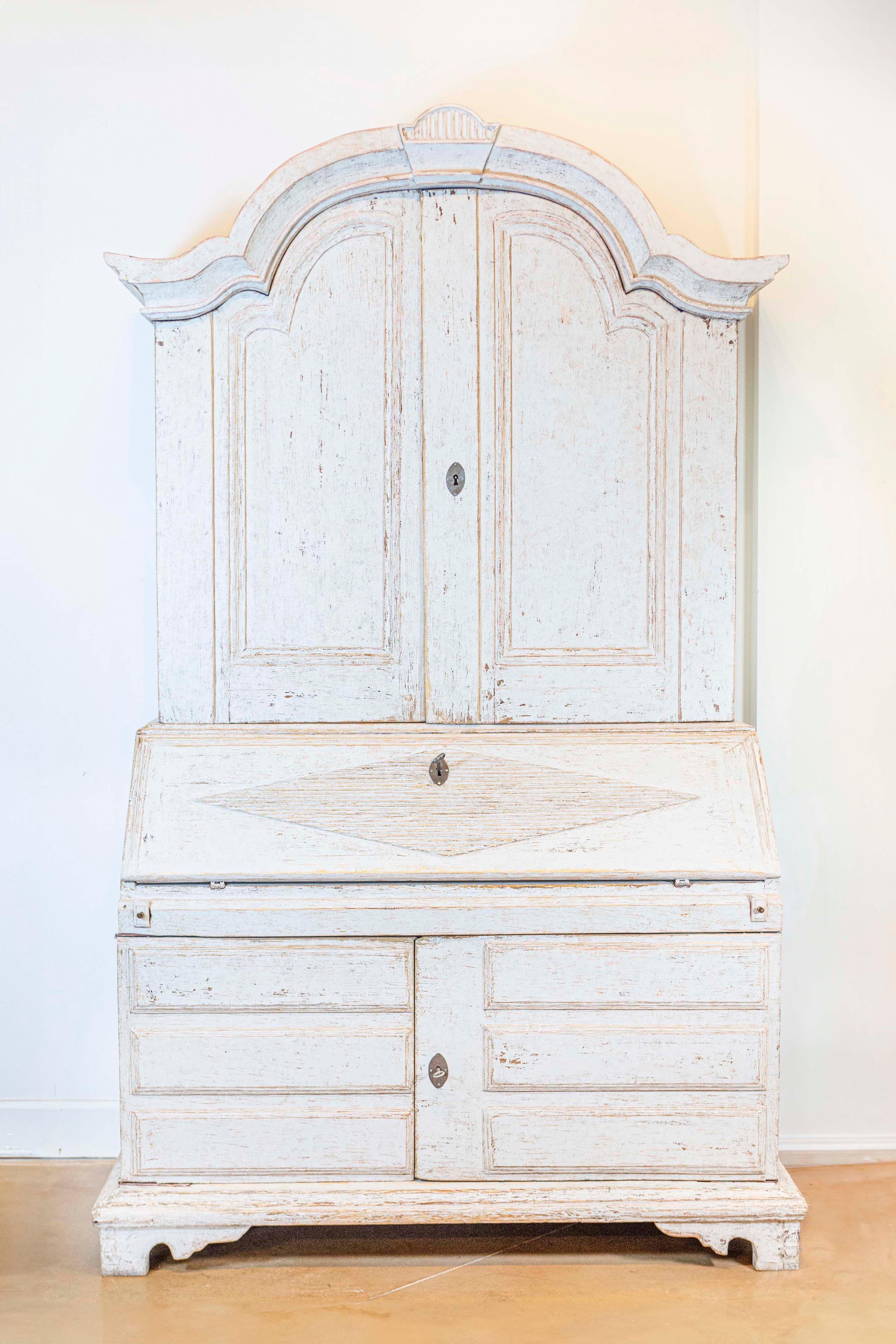 A Swedish tall secretary from Värmland circa 1800, with gray painted finish, carved bonnet top, four doors, slant front desk and bracket feet. Embrace the understated elegance of Swedish Gustavian design with this exquisite tall secretary from