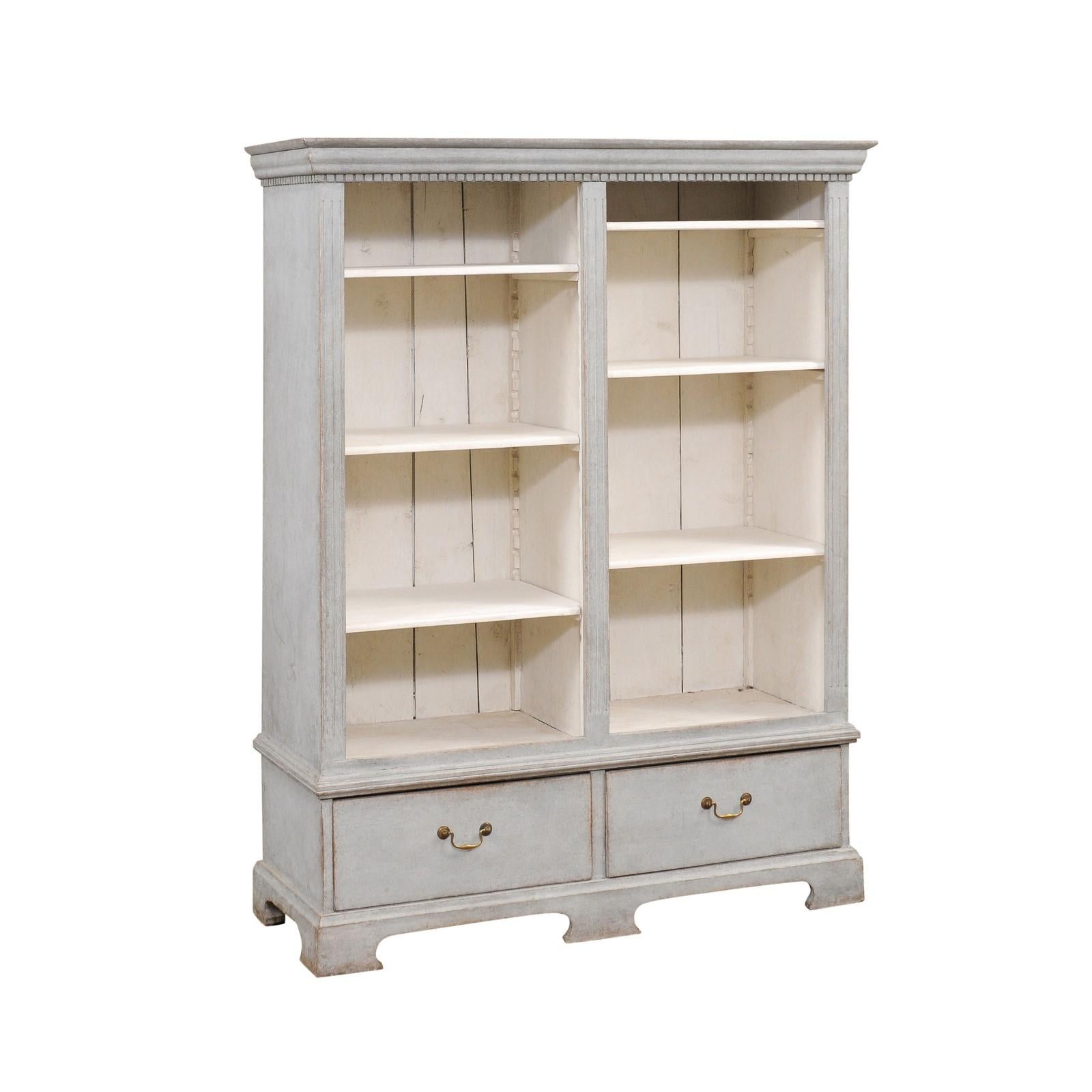 A Swedish wooden bookcase from circa 1850 with gray painted finish, carved dentil molding, open shelves, two drawers and bracket feet. This Swedish wooden bookcase from around 1850 stands as a testament to the timeless allure of antique