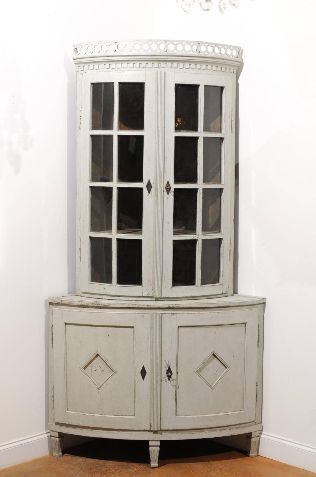 A Swedish Gustavian style painted corner cabinet from the mid-19th century with glass doors, pierced cornice and diamond motifs. This exquisite Swedish corner cabinet features a convex façade, adorned with a pierced cornice at the top, overhanging a