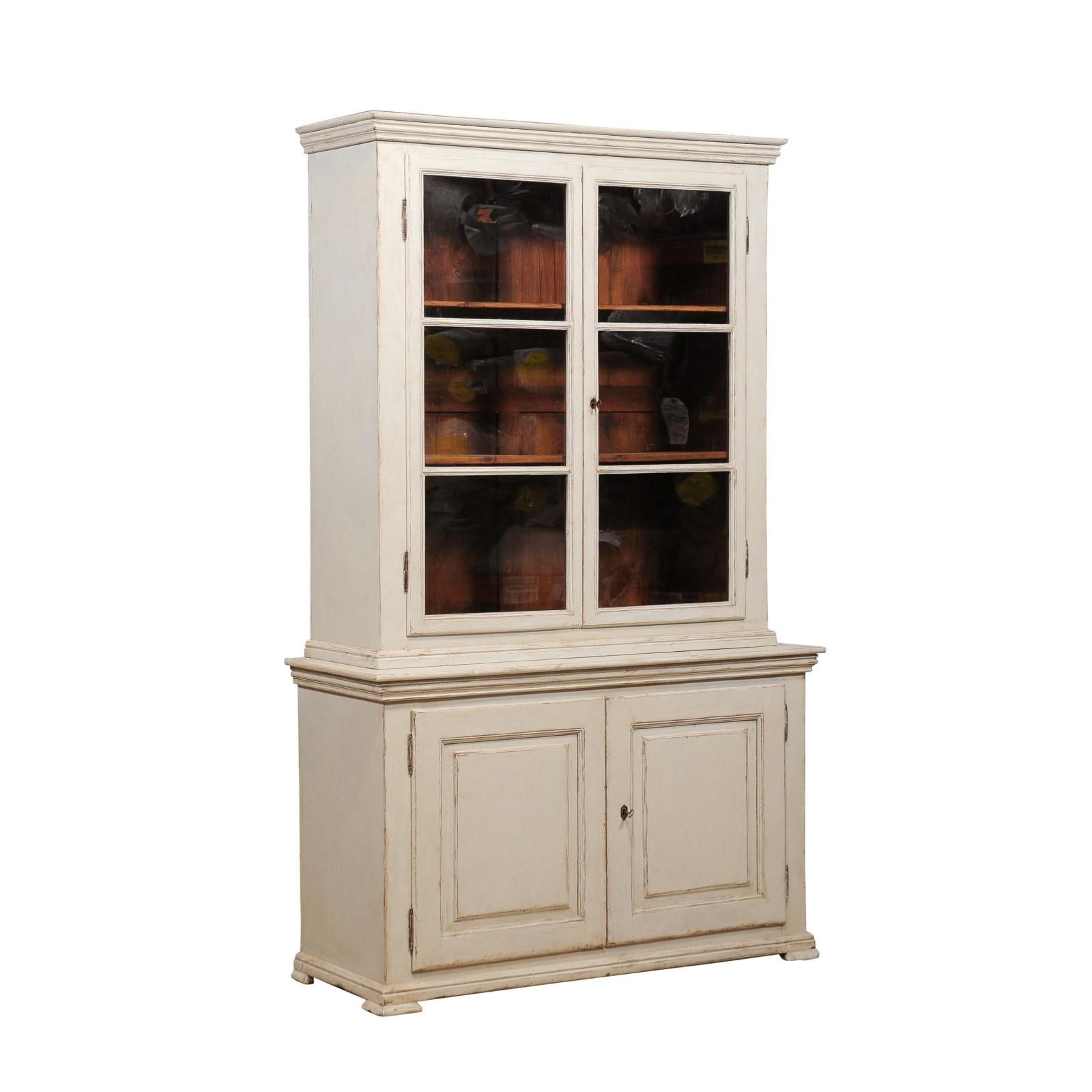 A Swedish two part glass door vitrine cabinet from circa 1850 with gray/beige painted finish and carved panels on the doors. This exquisite Swedish two-part glass door vitrine cabinet, crafted around 1850, effortlessly blends form and function. Its
