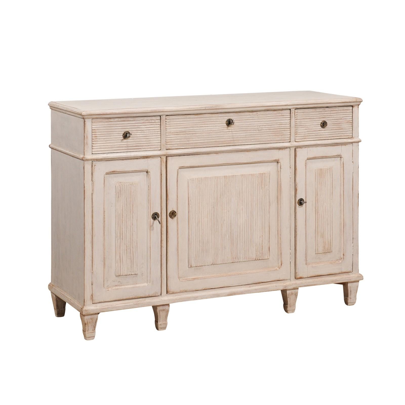 A Swedish Gustavian style sideboard from circa 1860 with light creamy gray painted finish, three drawers over three doors and carved reeded panels. This Swedish Gustavian style sideboard from circa 1860 exudes the elegance and refined simplicity