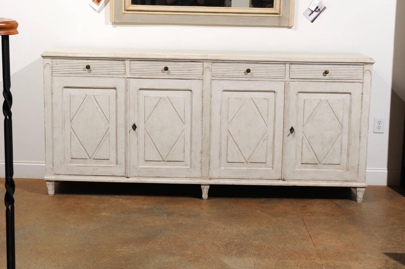 A Swedish Gustavian style painted sideboard from the late 19th century, with carved diamond patterns, reeded motifs and four drawers over four doors. This long Swedish painted sideboard features a rectangular top with molded edges, sitting above