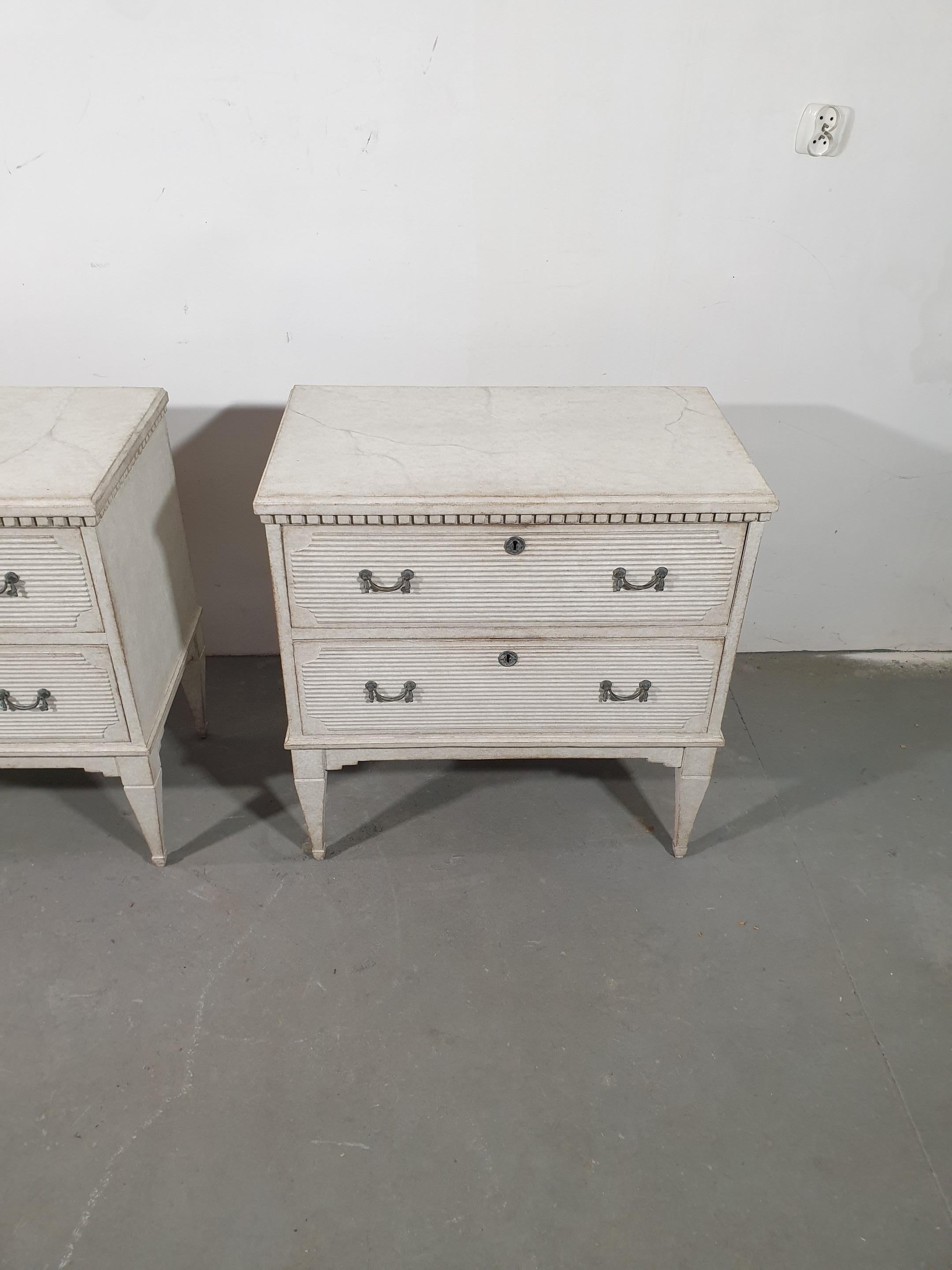 A pair of swedish Gustavian style chests from circa 1890 with rich light gray painted finish, carved dentil molding, two reeded drawers and tapered legs. Embrace the understated elegance of Swedish Gustavian design with this exquisite pair of chests