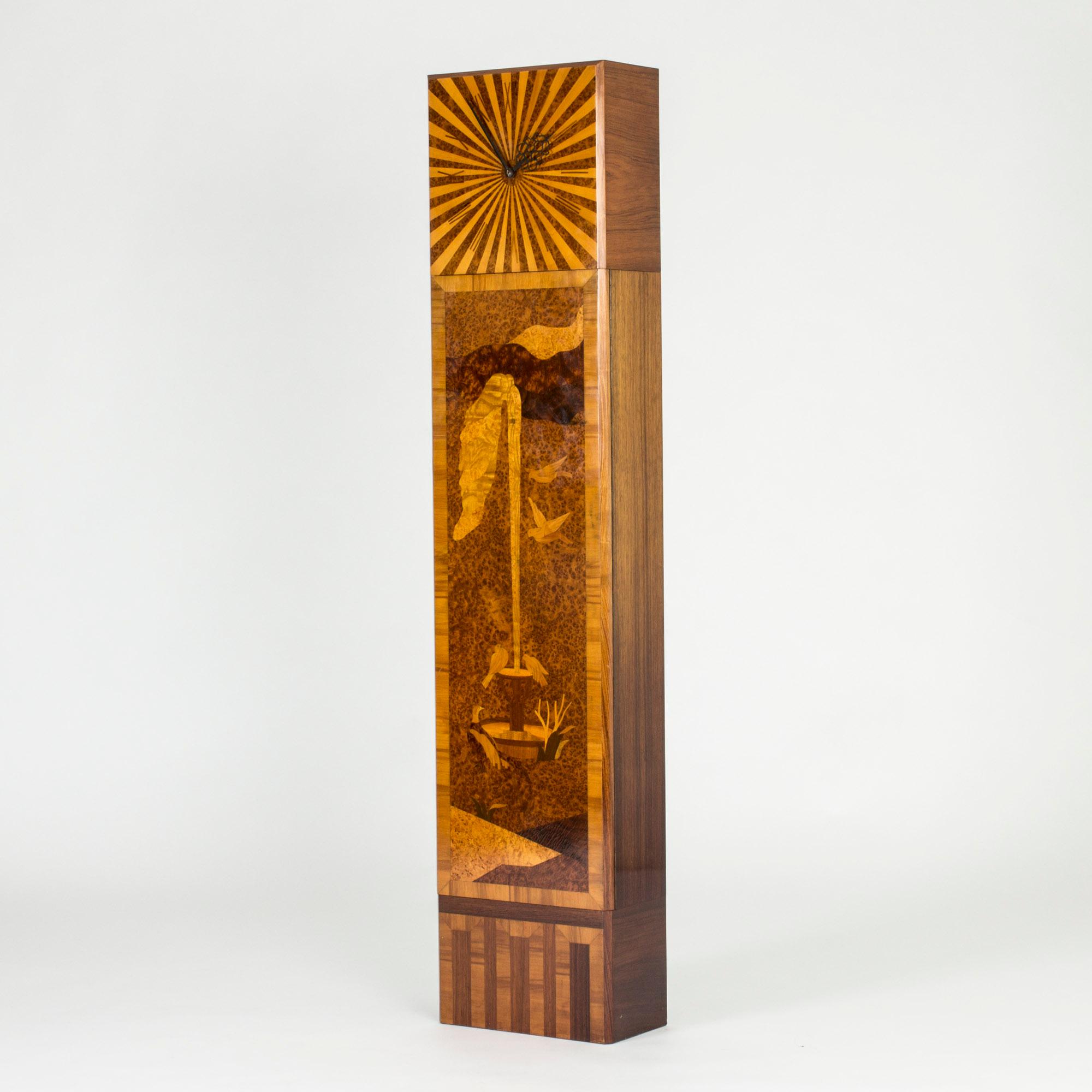 Striking 1930s floor clock from Mjölby Intarsia. Sleek, clean design and a beautiful inlayed motif of birds around a fountain. Beams spread out from behind the clockwork. The wood on the sides is layed with wood grain in contrasting directions.