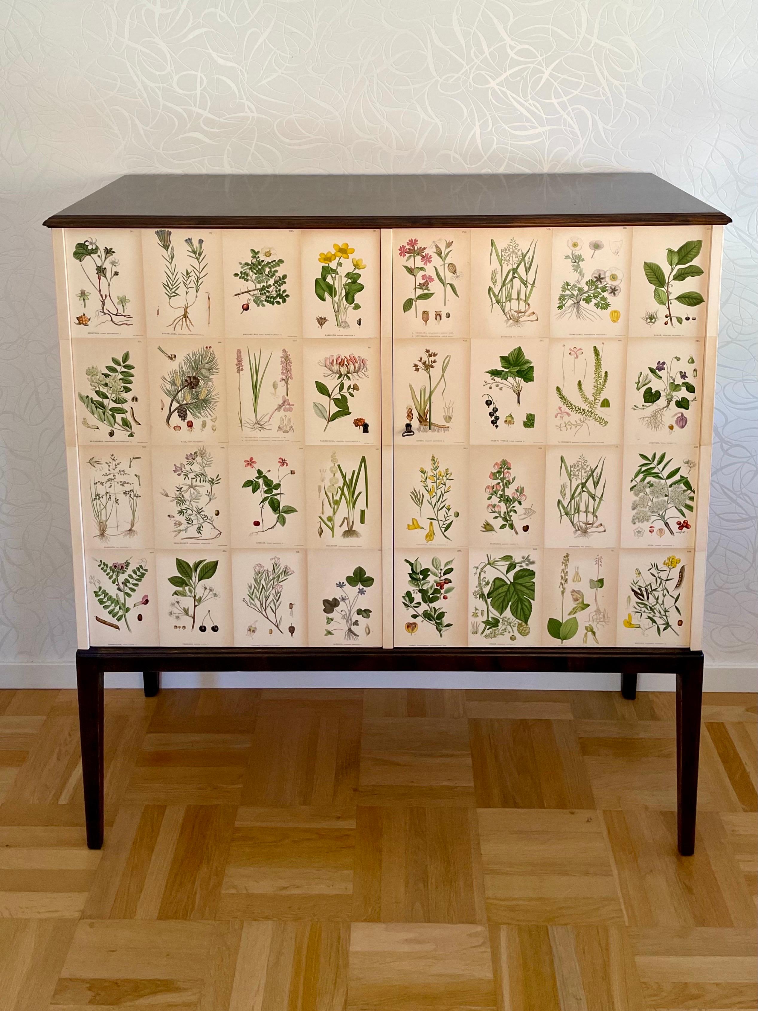 This is the Swedish Modern 1940s Cabinet, decorated with 56 different Nordens Flora-illustrations, in the style the famous head designer at Svenskt Tenn, Josef Frank.

This cabinet comes with a top veneered in dark stained birch.
It has later been