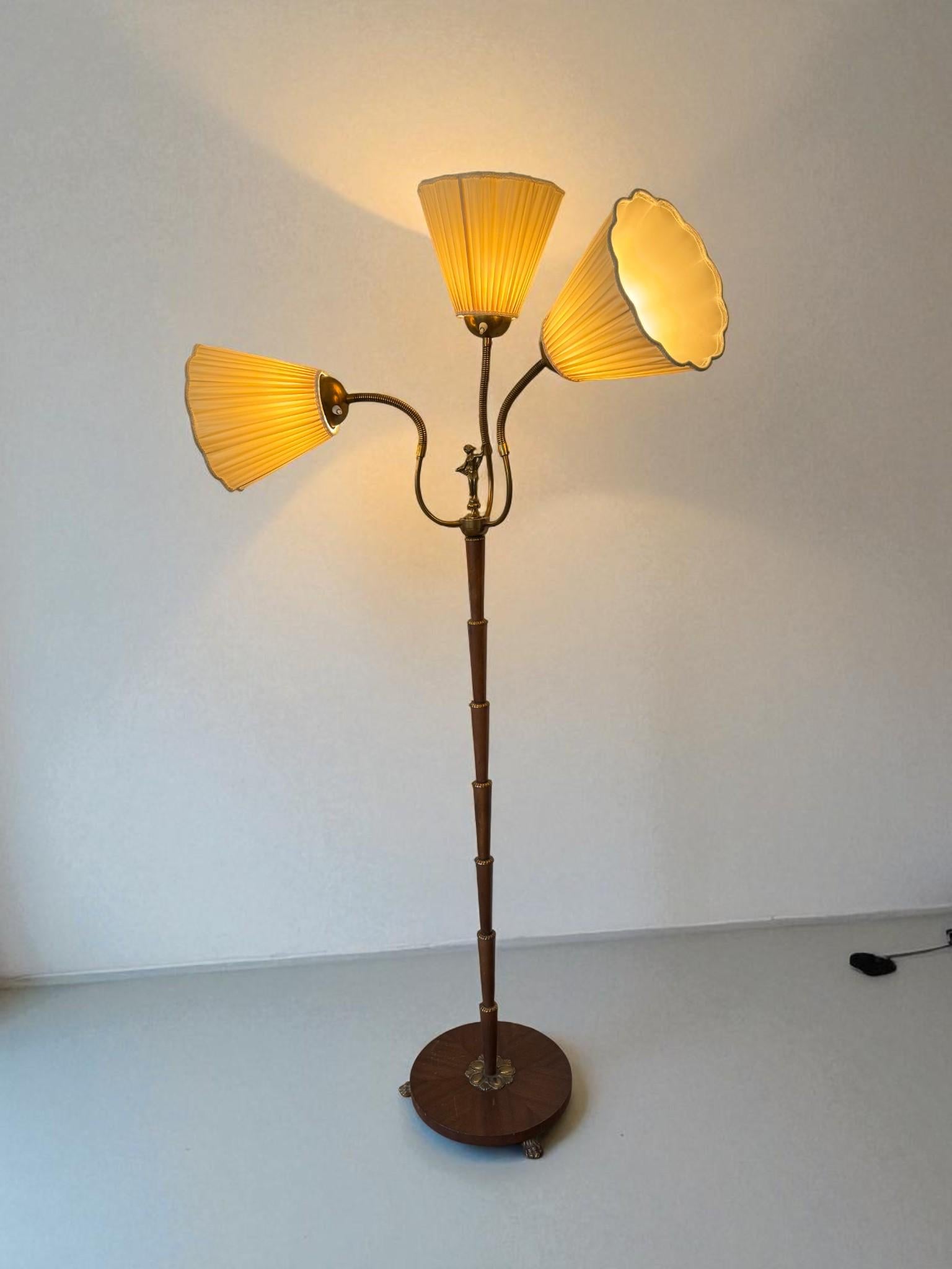 Encantadora lámpara de pie sueca de mediados de siglo, de los años 30. Esta elegante pieza presenta un tallo de madera finamente segmentado que se eleva desde una base circular, con tres brazos de latón graciosamente curvados que sostienen pantallas