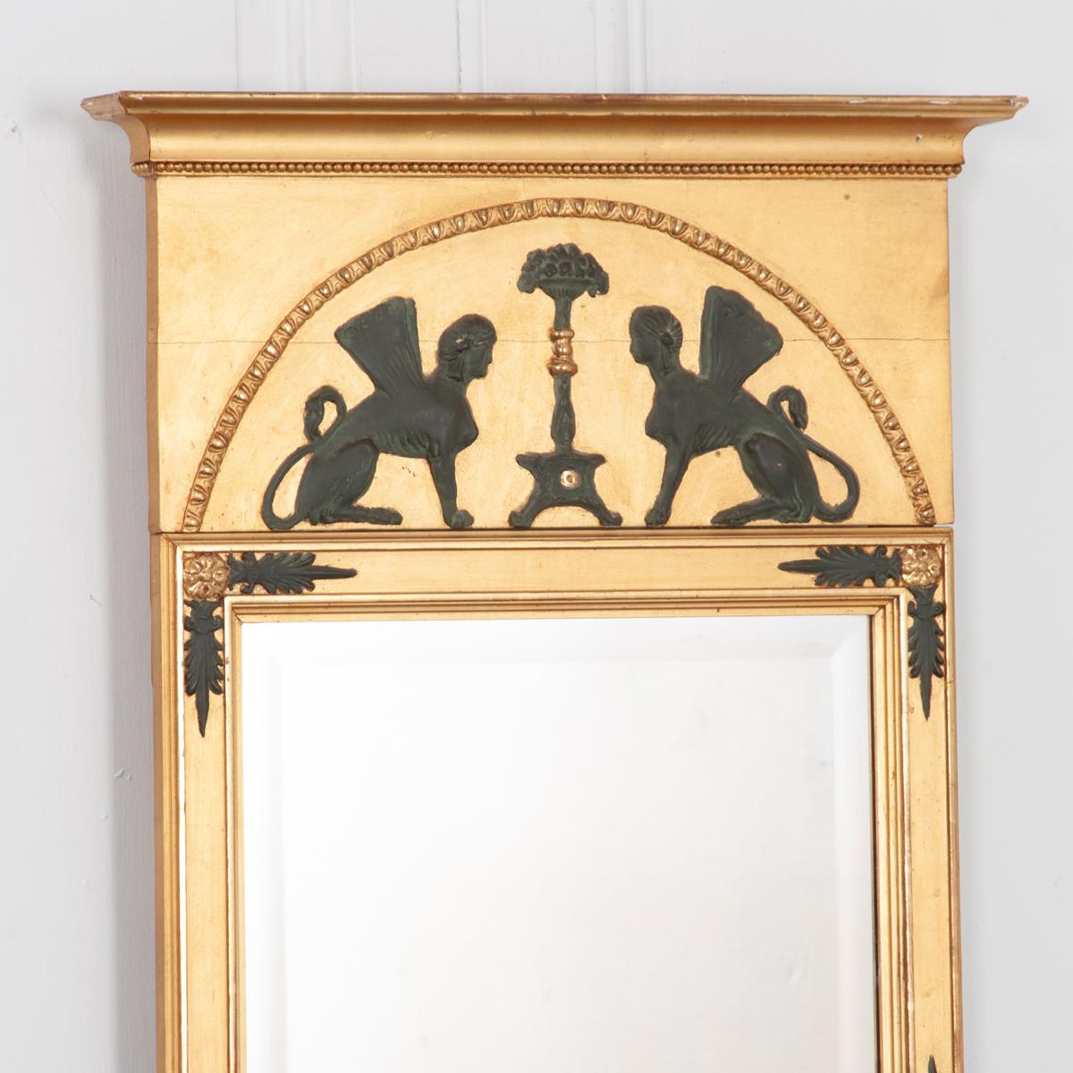 This mirror is an exceptional example of an Empire style trumeau from Sweden, circa 1850 with gorgeously carved Egyptian inspired details. The beveled mirror glass is framed in gold gilt with painted black floral details. The whole is adorned with