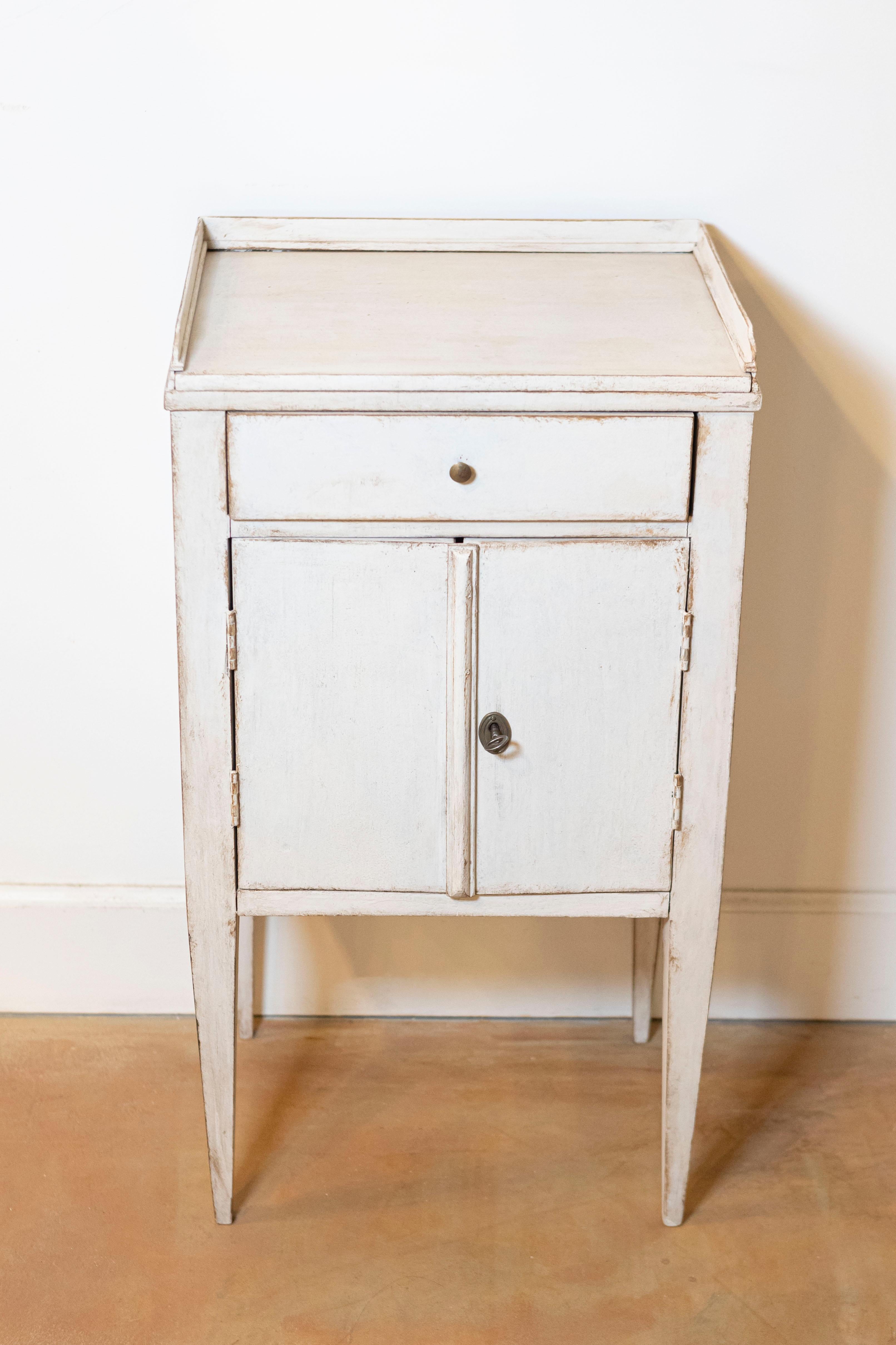 A Swedish bedside table from the 19th century with gray/cream painted finish, three-quarter gallery encircling the top, single drawer, petite double doors and tapered legs. This 19th-century Swedish bedside table is a charming piece that beautifully