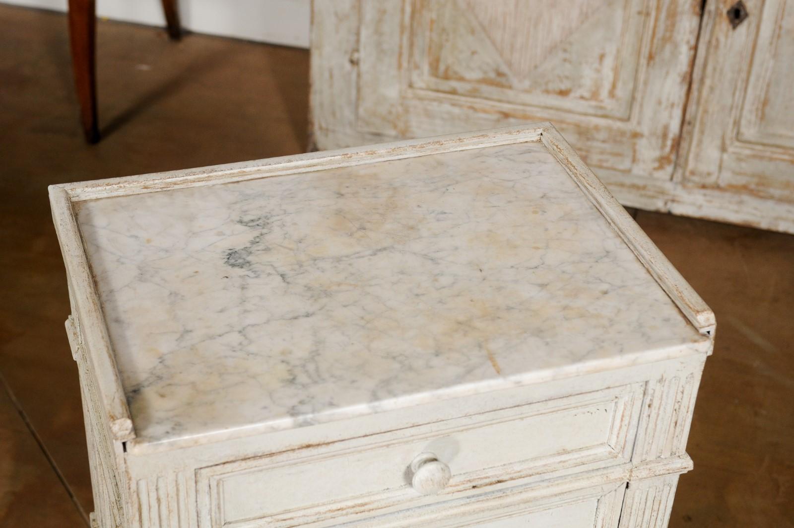 A Swedish neoclassical style painted wood nightstand table from the 19th century, with marble top, drawer, door and fluted motifs. Born in Sweden during the 19th century, this elegant bedside table features a rectangular marble top with slightly