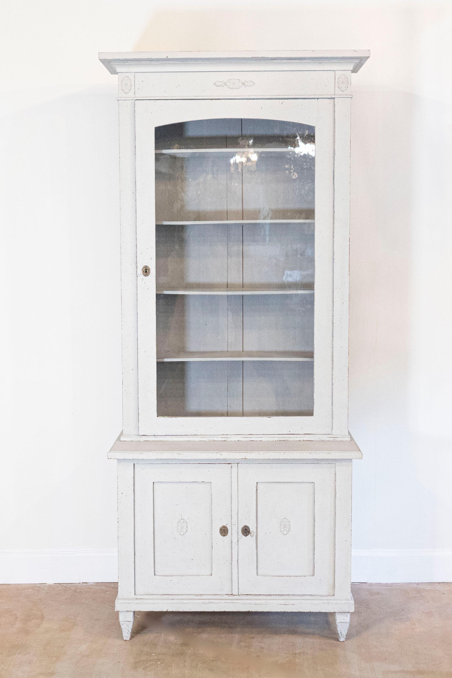 A Swedish Gustavian style painted wood vitrine from the 19th century, with carved rosettes. Created in Sweden during the 19th century, this vitrine cabinet features a beveled cornice sitting above a single glass door, topped with a carved floral