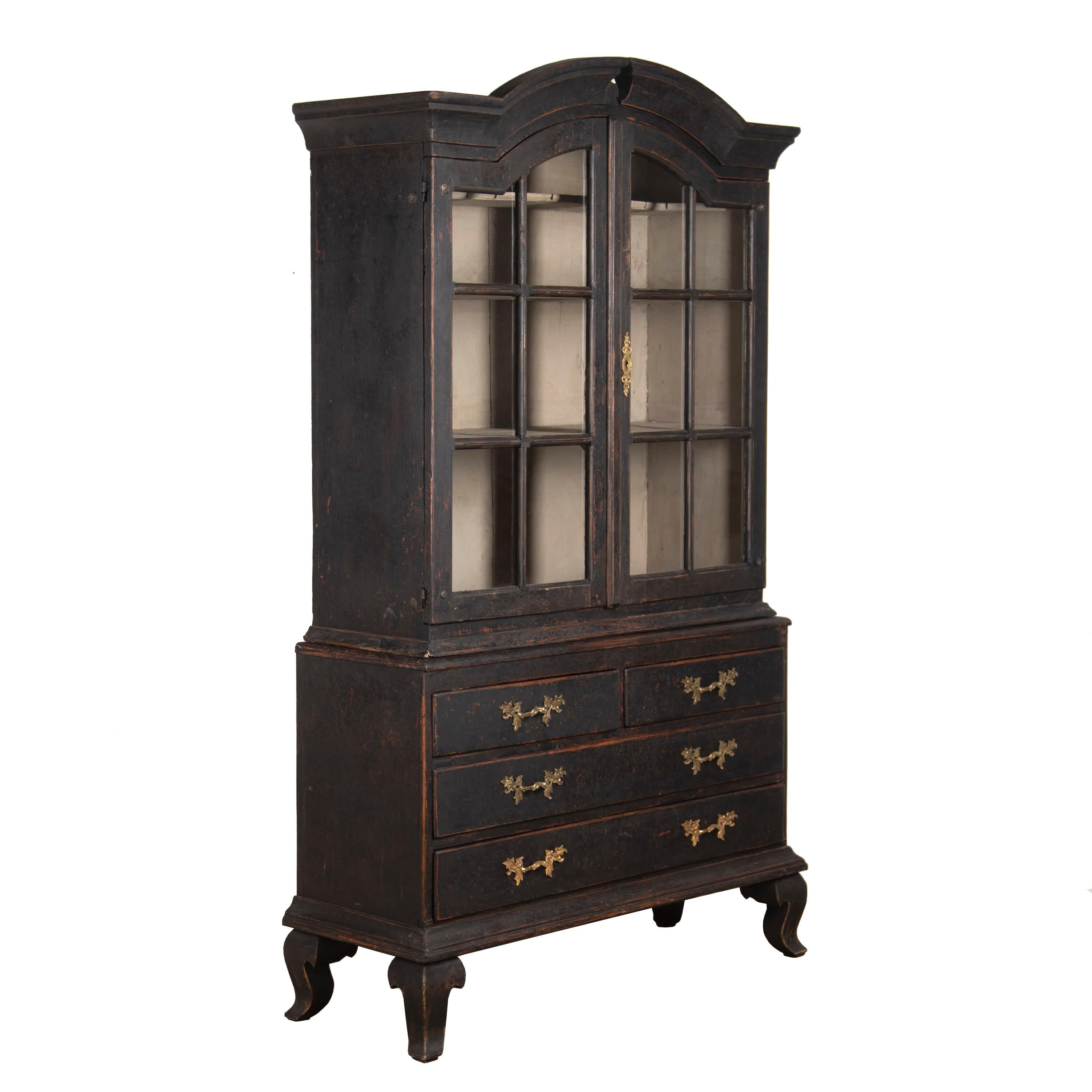 Swedish 19th century vitrine with a decorative carved wooden pediment and tapering to cabriolet legs. Glazed doors with original glass open to display shelves, and below are two short and two long drawers. This piece has been repainted in black,