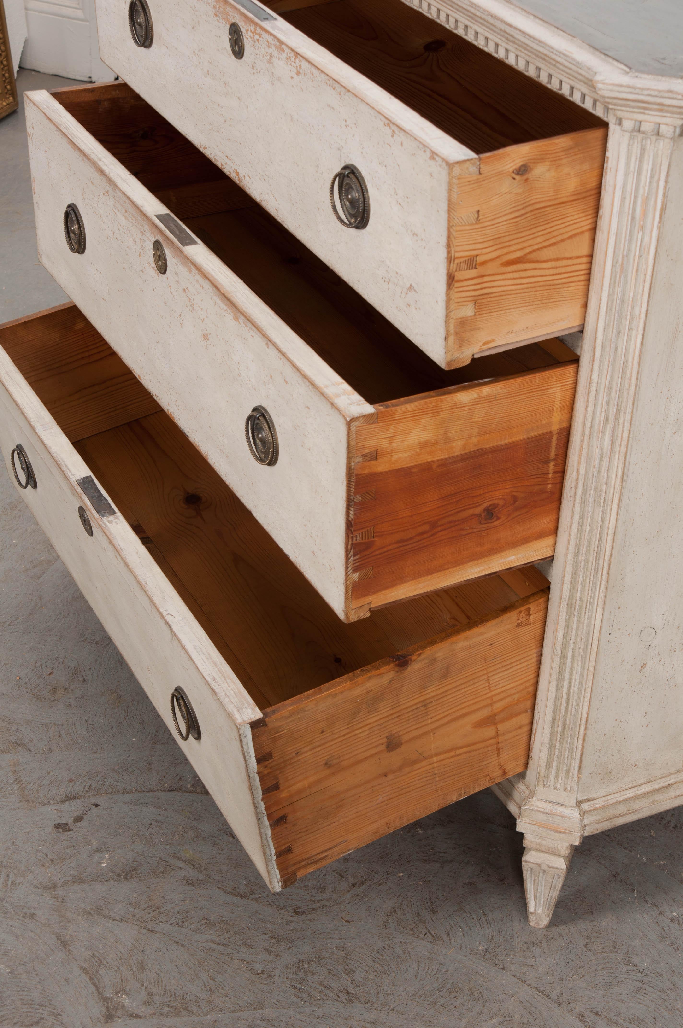 This lovely Swedish neoclassical-style chest, circa 1900, is sure to brighten up any space! Featuring a partially painted conforming surface with dentillated molding over canted and fluted corners and a bank of three graduated drawers, each with