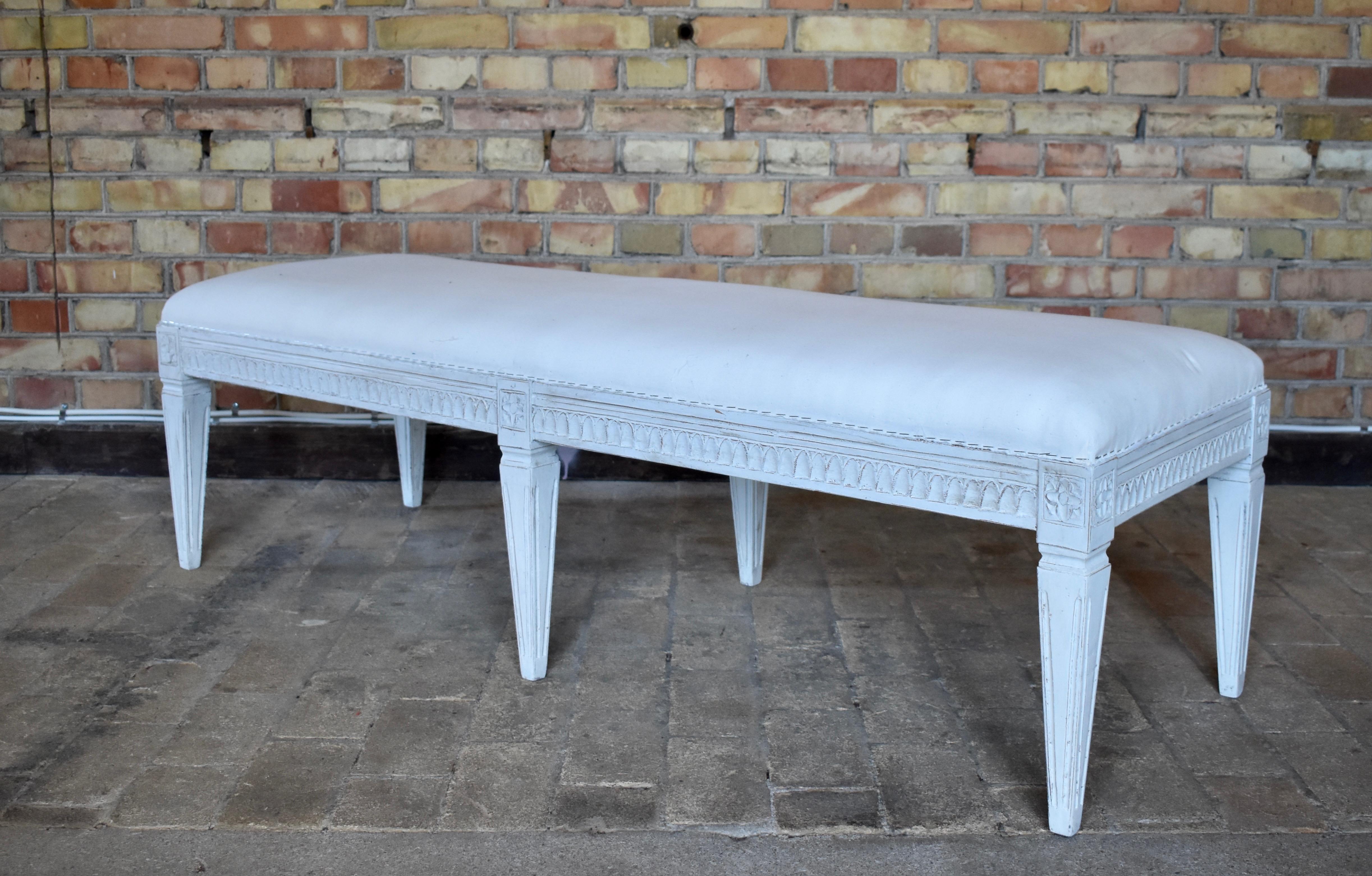 Antique Swedish Gustavian painted bench, with carved lamb's tongue apron, square tapered legs with carved flower rosettes at top of each leg. The bench is painted in a white-greyish color and distressed finish. Renewed upholstery and textile.