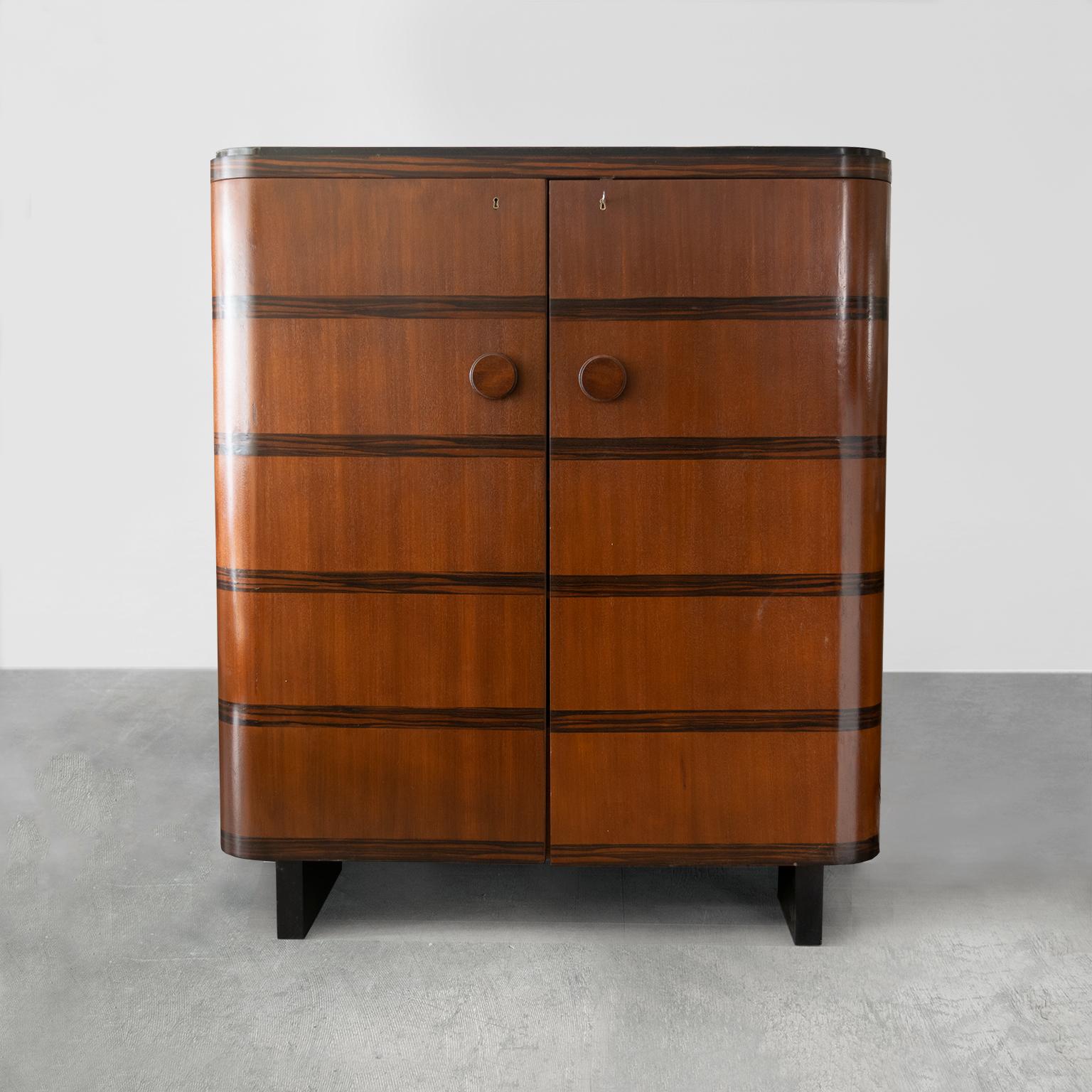 A large Swedish Art Deco 2-door cabinet veneered in Mahogany with horizontal bands of Palisander Ebony. The cabinet has 4 adjustable shelves and 2 spacious built-in drawers. The front curved doors follow the cabinets streamlined form and 2 black