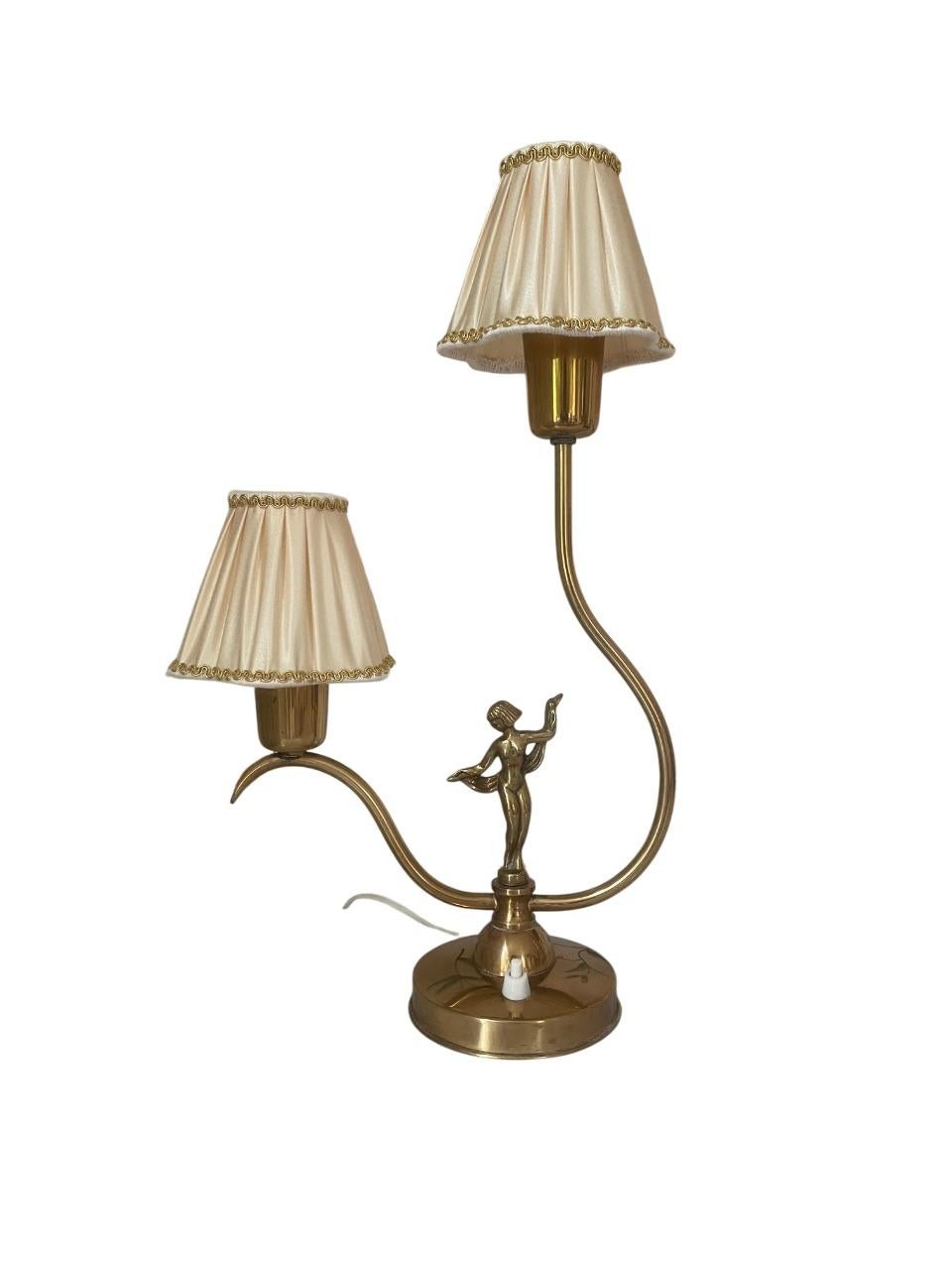 Charmante et sculpturale lampe de table en laiton d'Ystad-Metall, Suède, datant des années 1930. Cette pièce élégante présente un design distinctif à double bras, avec deux courbes enveloppantes supportant des abat-jour en tissu. Au centre se trouve
