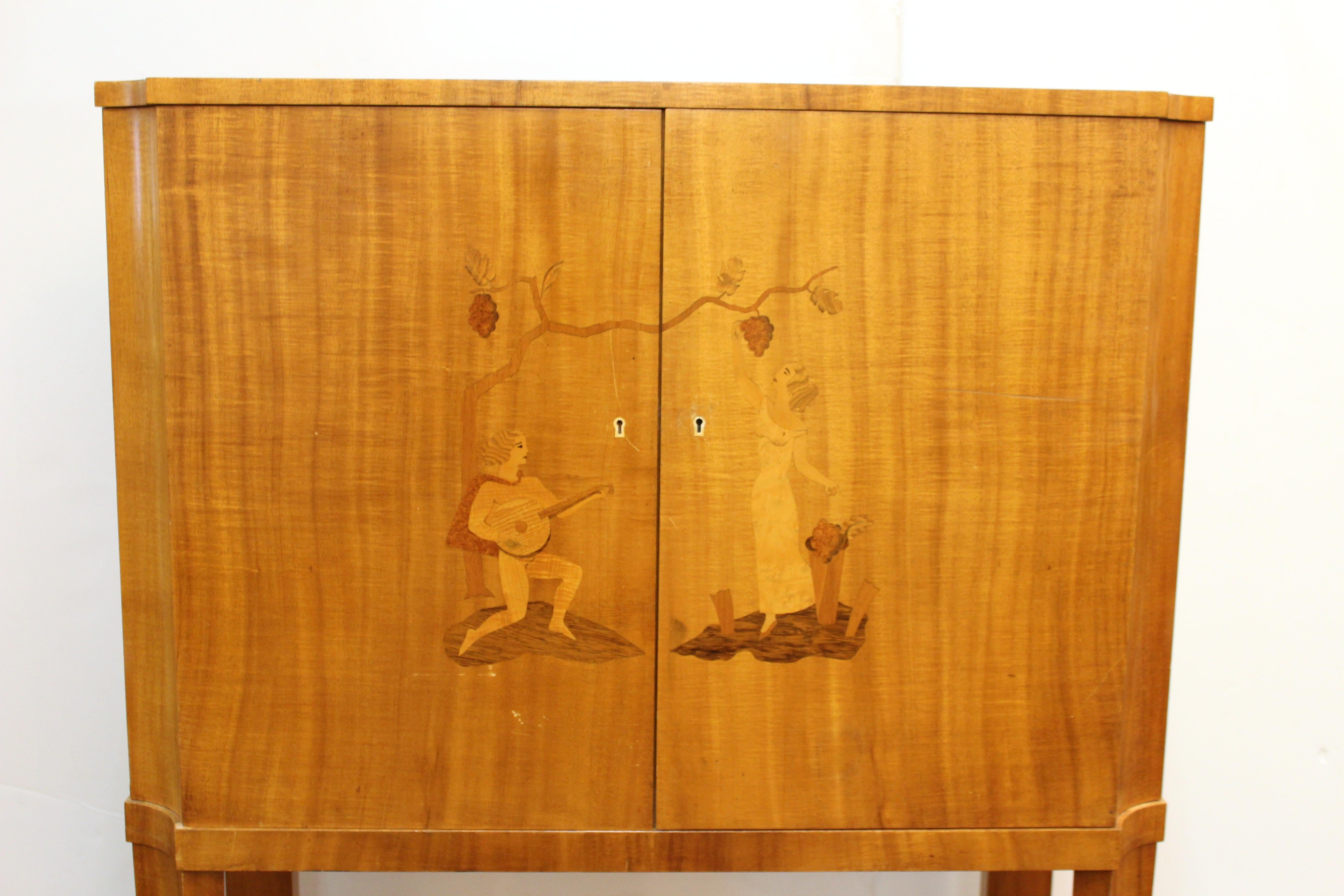 Art Deco bar cabinet from Mjolby, Sweden in blond wood with inlaid illustration. The front depicts a young couple among grape vines including a man playing an instrument and a woman reaching up to inspect grapes on a vine above. The piece opens to