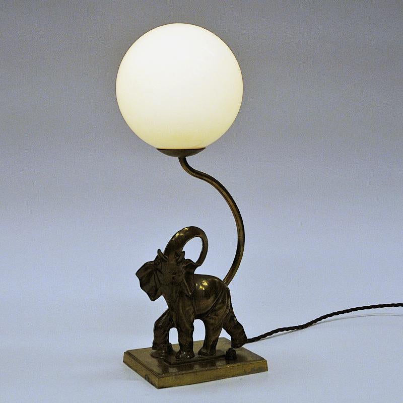 Swedish Art Deco table lamp with an off white opaline glass dome and brass elephant lamp base from the 1930s. The lamp has a round shiny dome lifted up by a brass pole companied with a walking elephant with its trunk lifted high. Black light switch