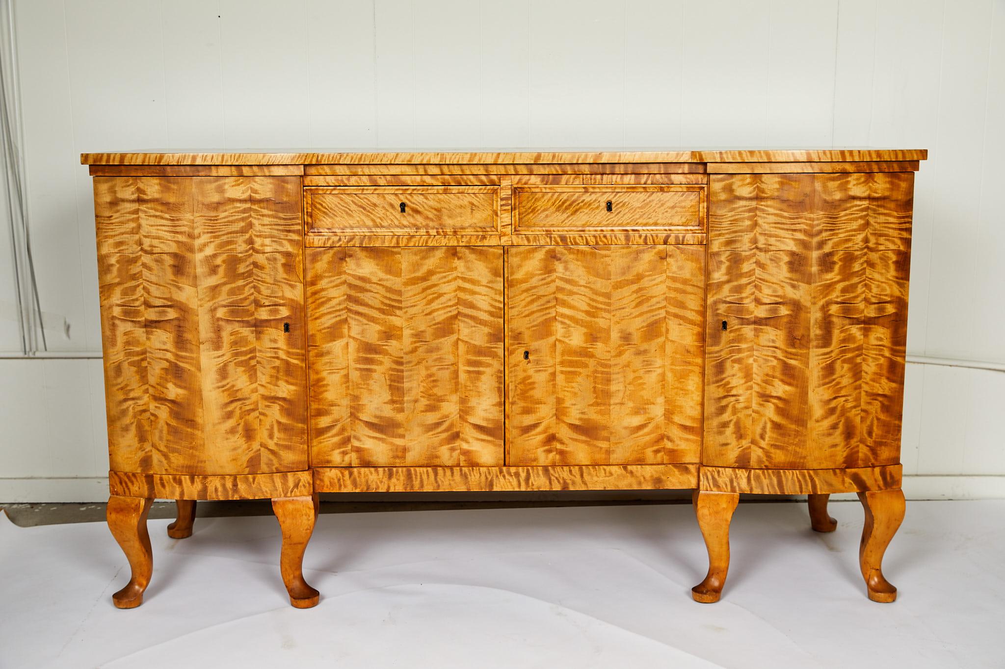 Early 20th century Swedish Art Deco period sideboard/cabinet rendered in highly figured, bookmatched golden flame birch. A shaped one board top is supported by a case holding two drawers and one center cabinet flanked by two outer cabinets. The case