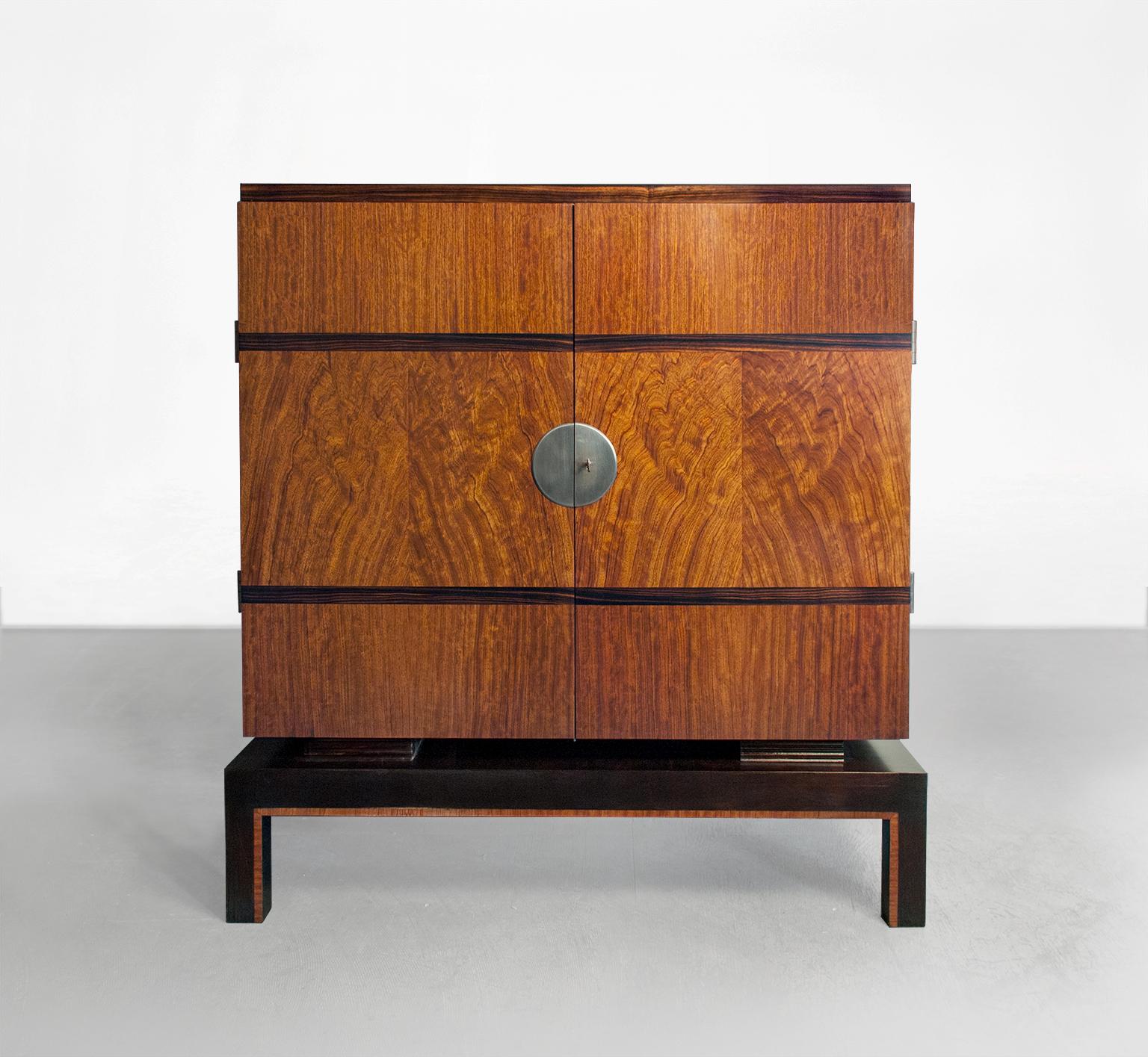 Swedish Art Deco two-door cabinet which hovers on a plinth and is veneered with mahogany and horizontal bands of Macassar ebony. A decorative steel circular plate covers the lock mechanism and exterior tubular steel hinges detail the cabinets sides.