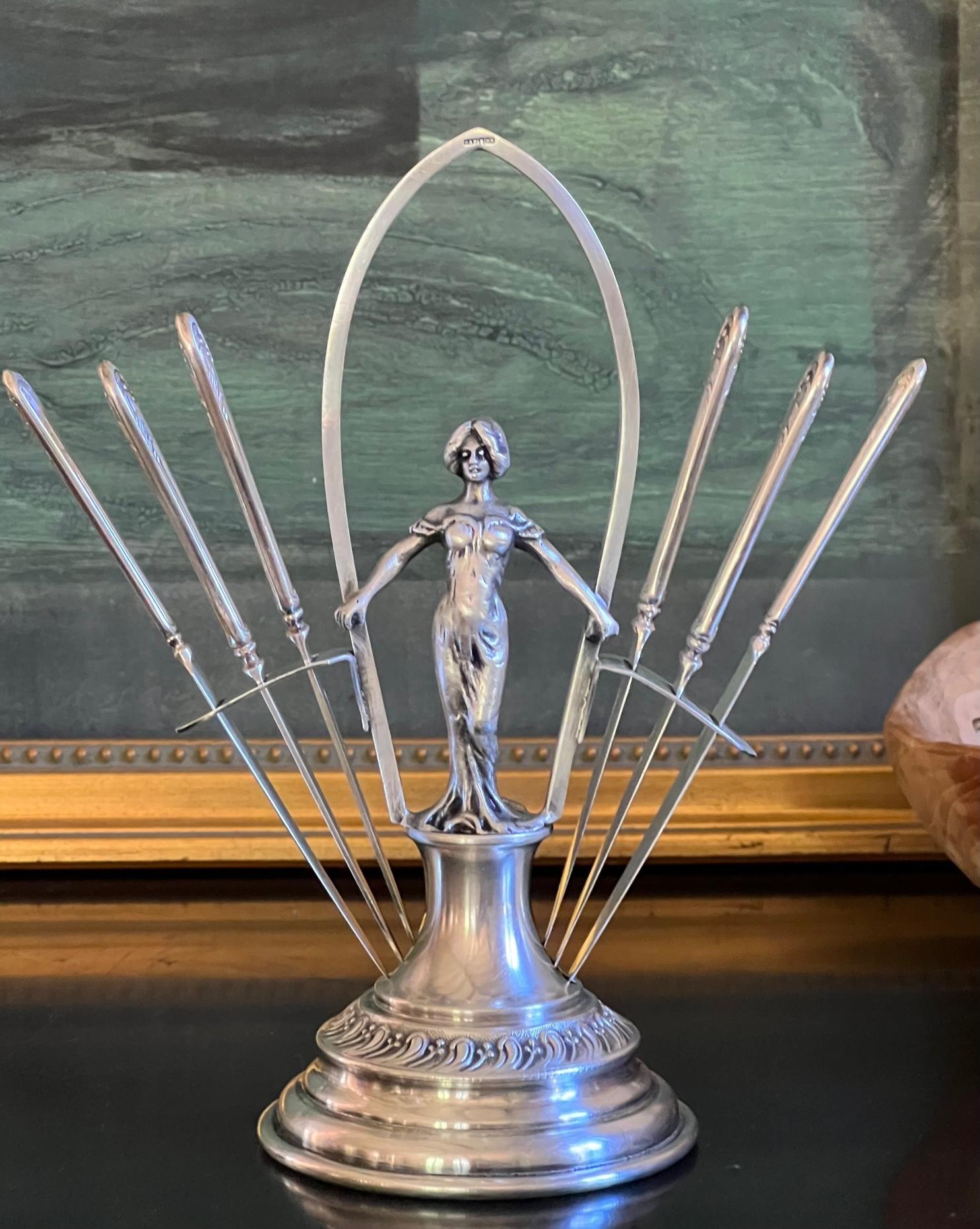 Art Nouveau set of fruit knives made by GAB (Guldsmeds Aktie Bolet) in Sweden sometime around 1900. The stand is a figure of a woman with her arms outstretched with three knives on each side.

Stamped on the tip of the stand is  GAB  MS
Each knife