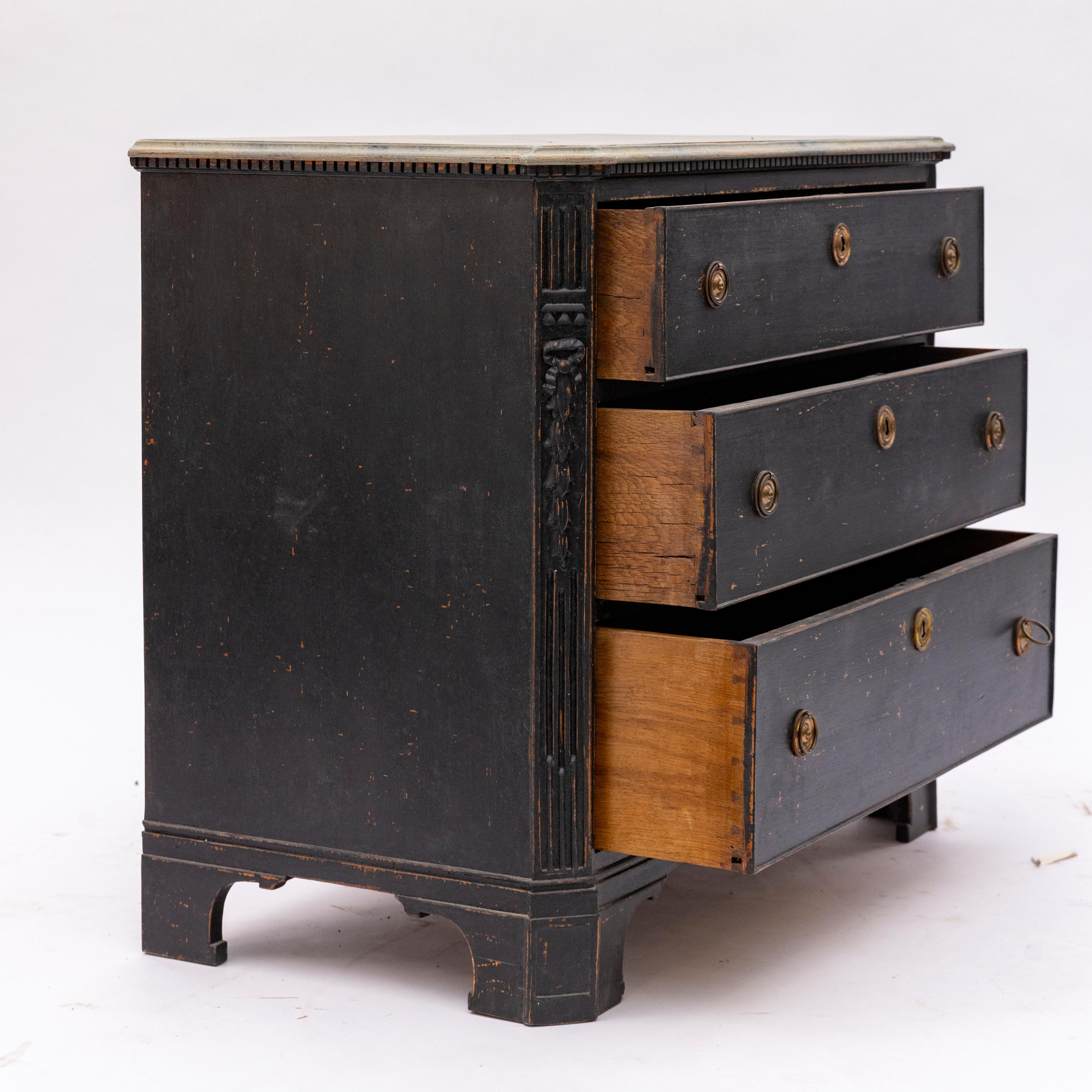 A black-painted Gustavian chest of drawers with three drawers, each adorned with patinated brass hardware.
The chest showcases elegant canted corners with fluted details and is crowned by a finely carved bow with foliage motifs.
The top is  finished